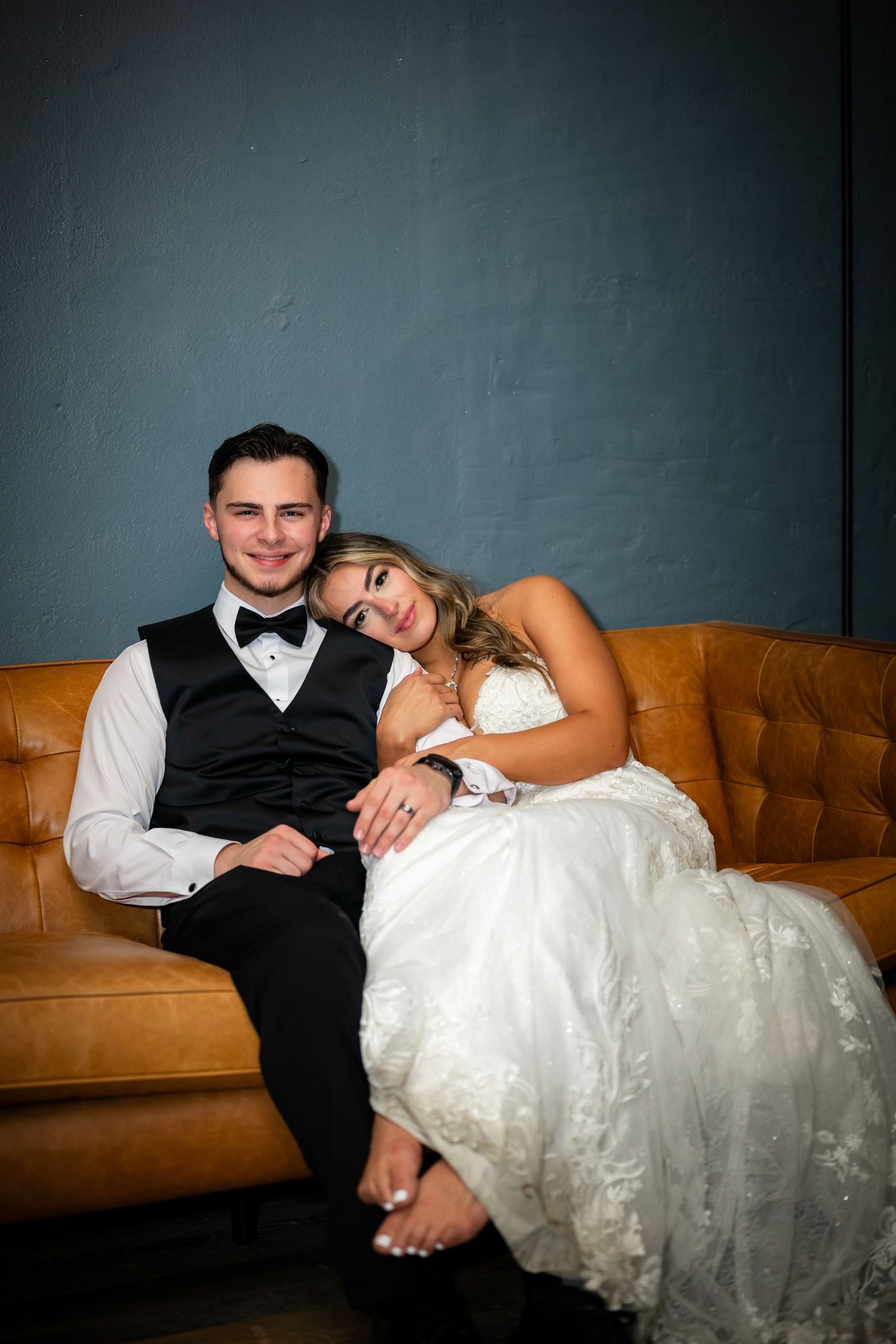 A couple relaxes on a brown leather couch against a dark wall, with the bride in a white lace gown resting on the groom.