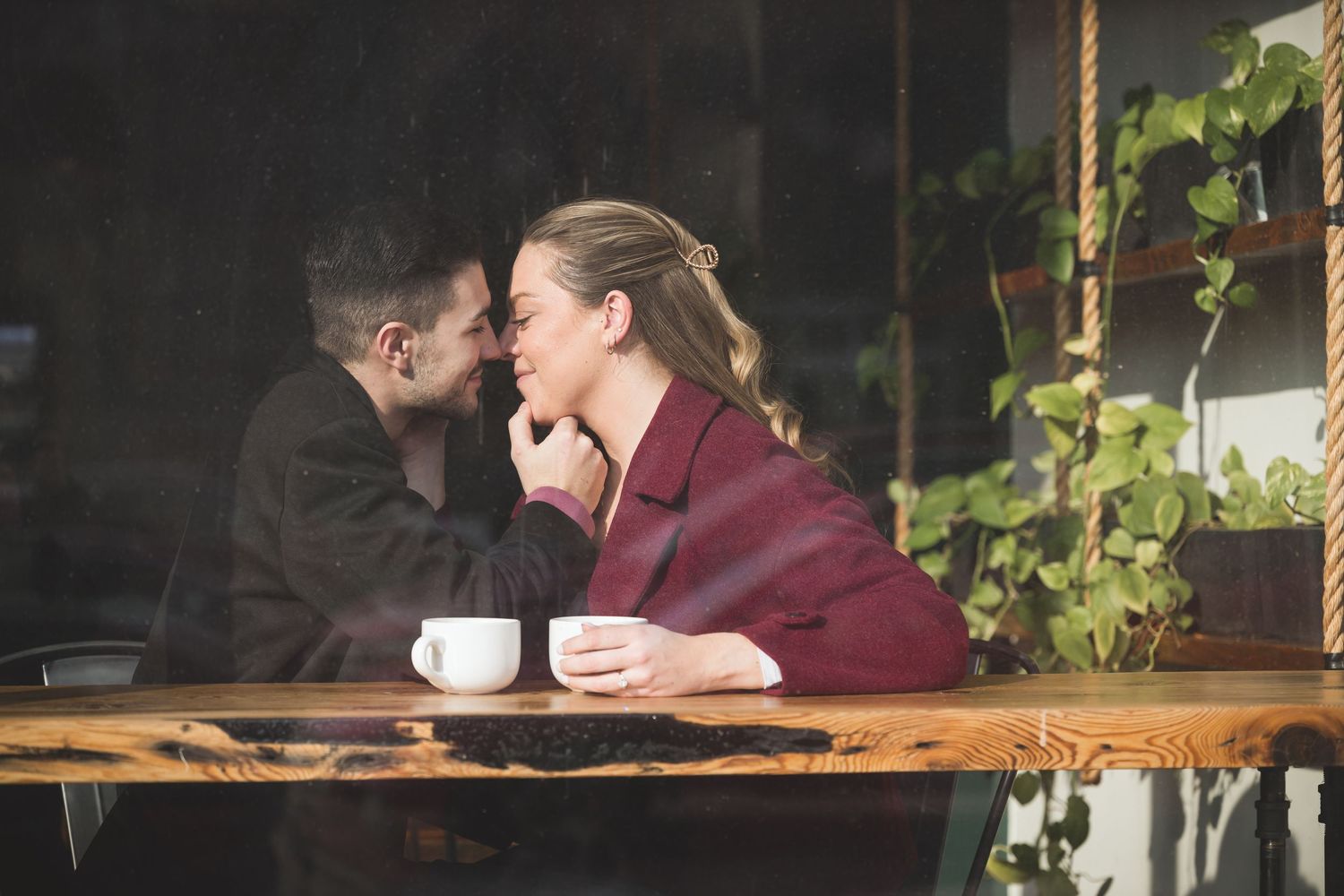Two people share an intimate conversation at a coffee shop table with greenery visible through the window.