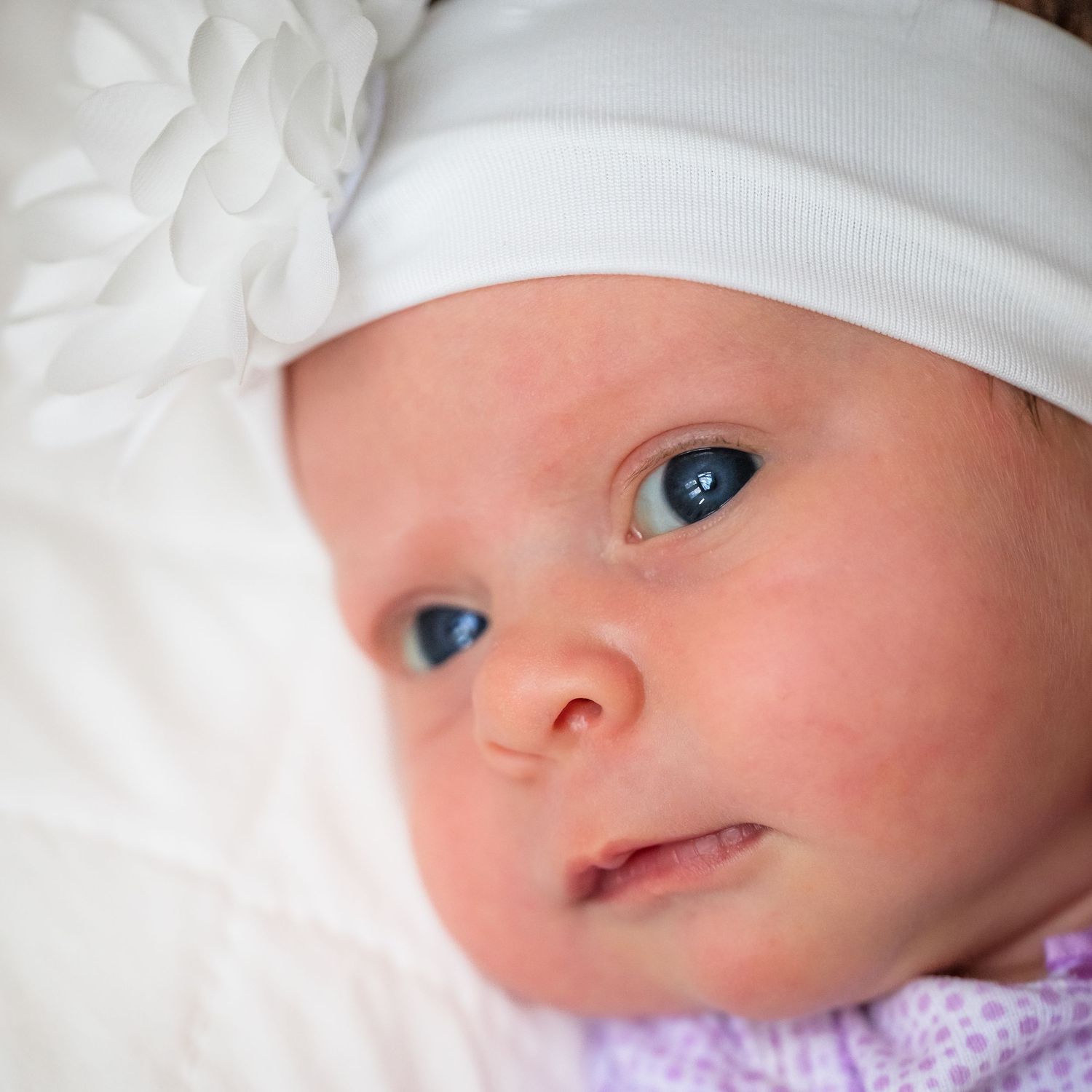 Close up of an infant wearing a white cap with bright blue eyes looking at the camera.