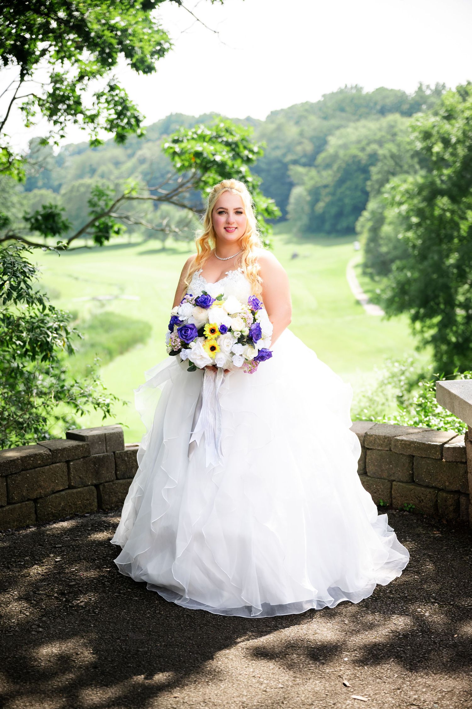 A bride in a full ballgown poses with purple and white flowers against a scenic hillside backdrop.