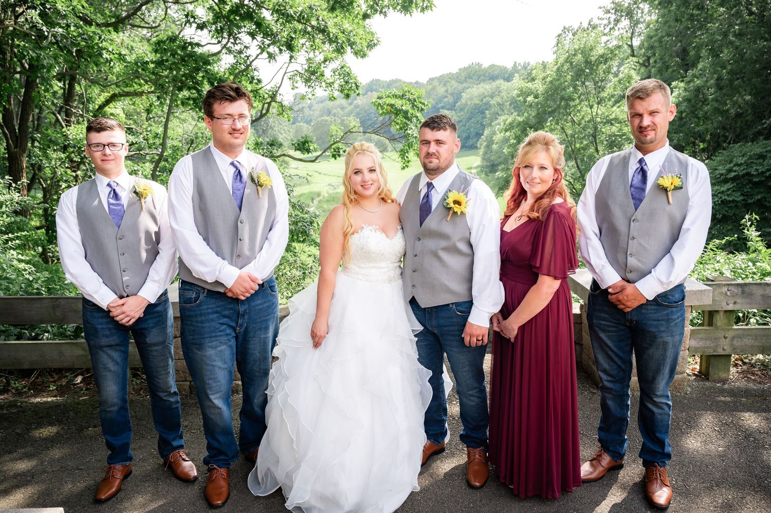 Wedding party in gray vests and purple gowns poses outdoors on a stone terrace surrounded by lush greenery.