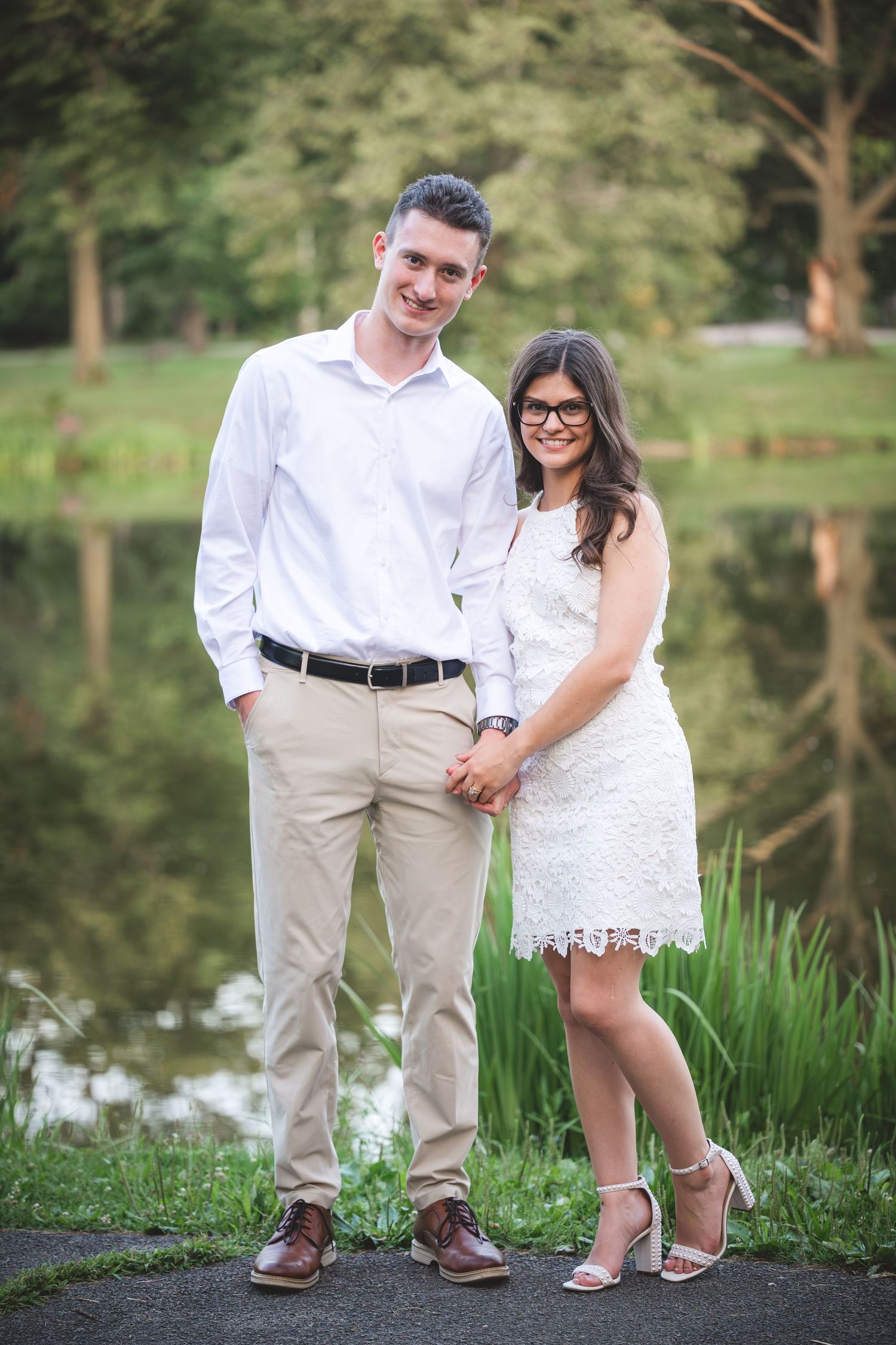 Couple holding hands in white outfits posing by a serene pond surrounded by lush greenery.
