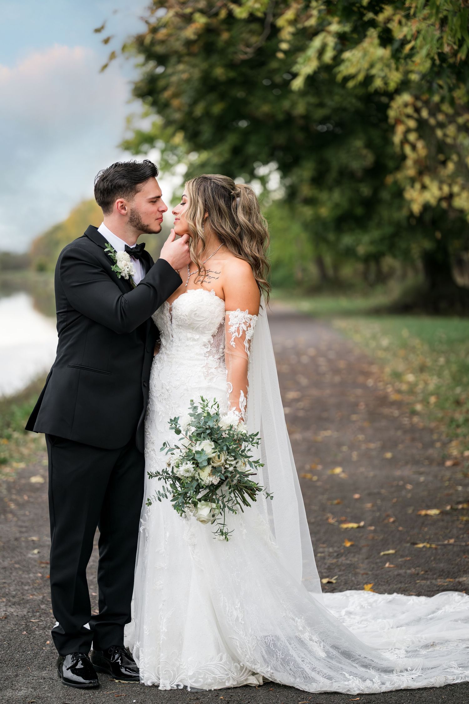 A romantic wedding couple shares tender moments on a path lined with autumn trees in their formal attire.