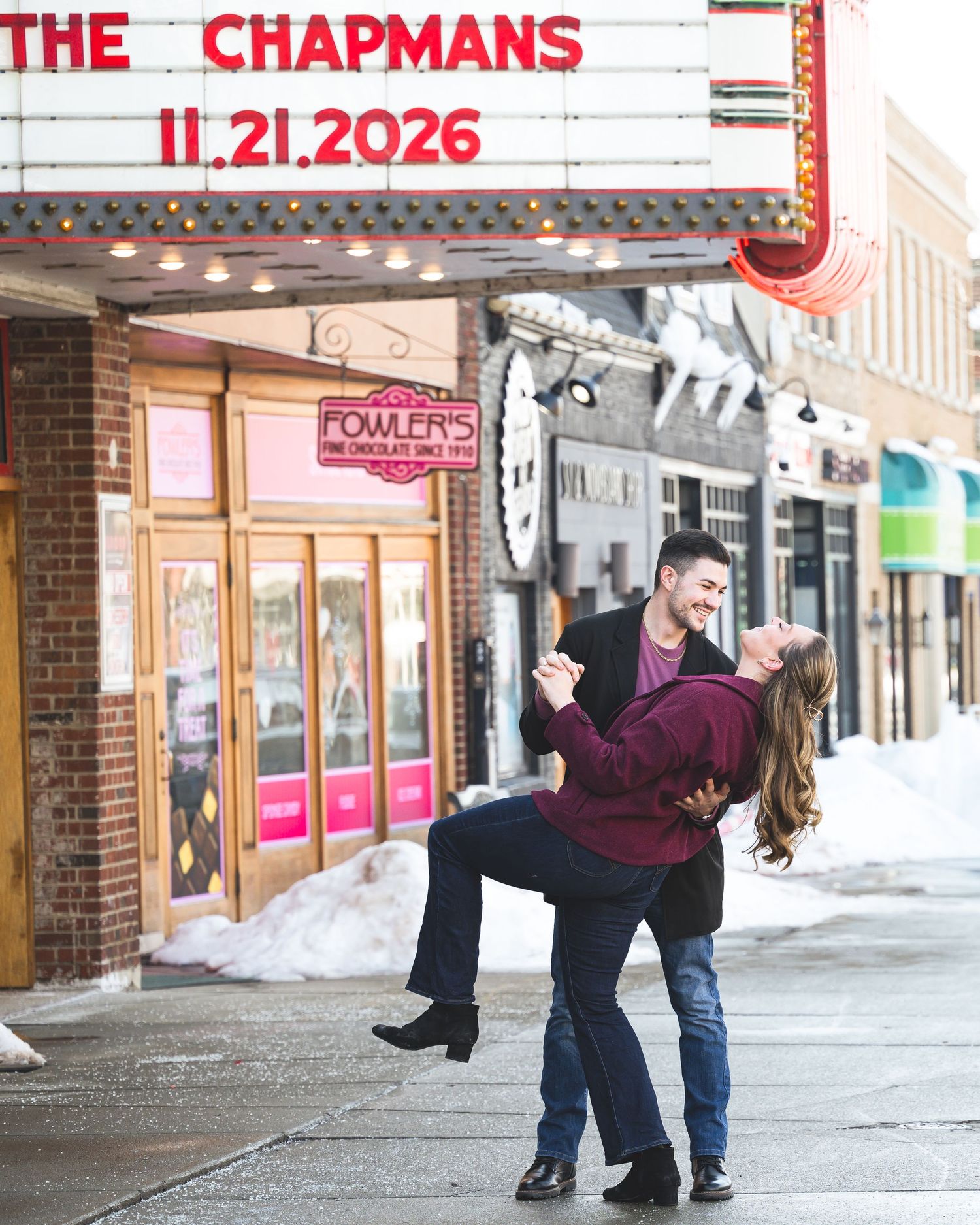 Couple dancing under a theater marquee announcing The Chapmans wedding date 11.21.2026 in a snowy downtown street.