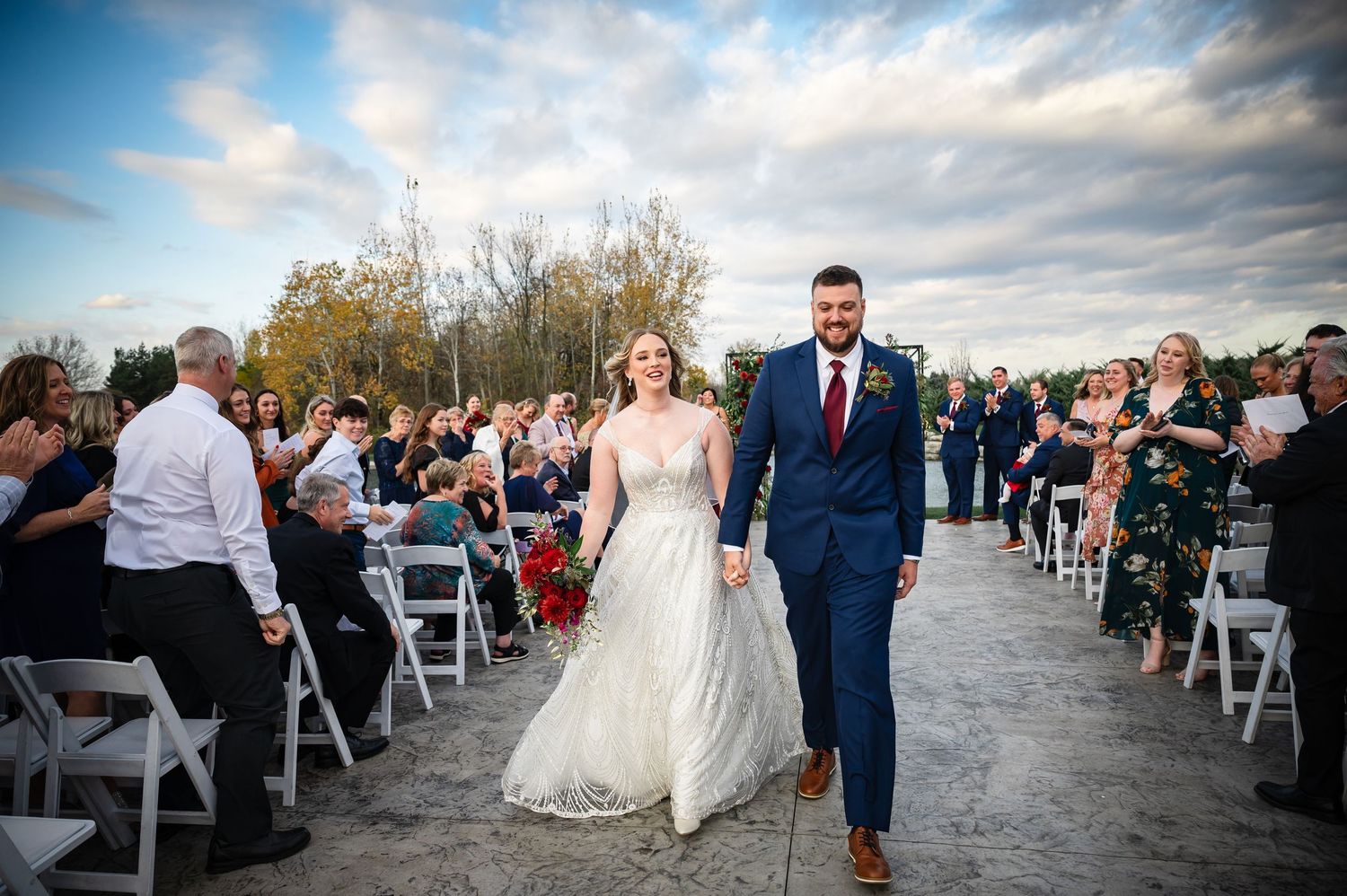 Outdoor wedding celebration under blue skies with guests seated in rows of white chairs beside a waterfront location.
