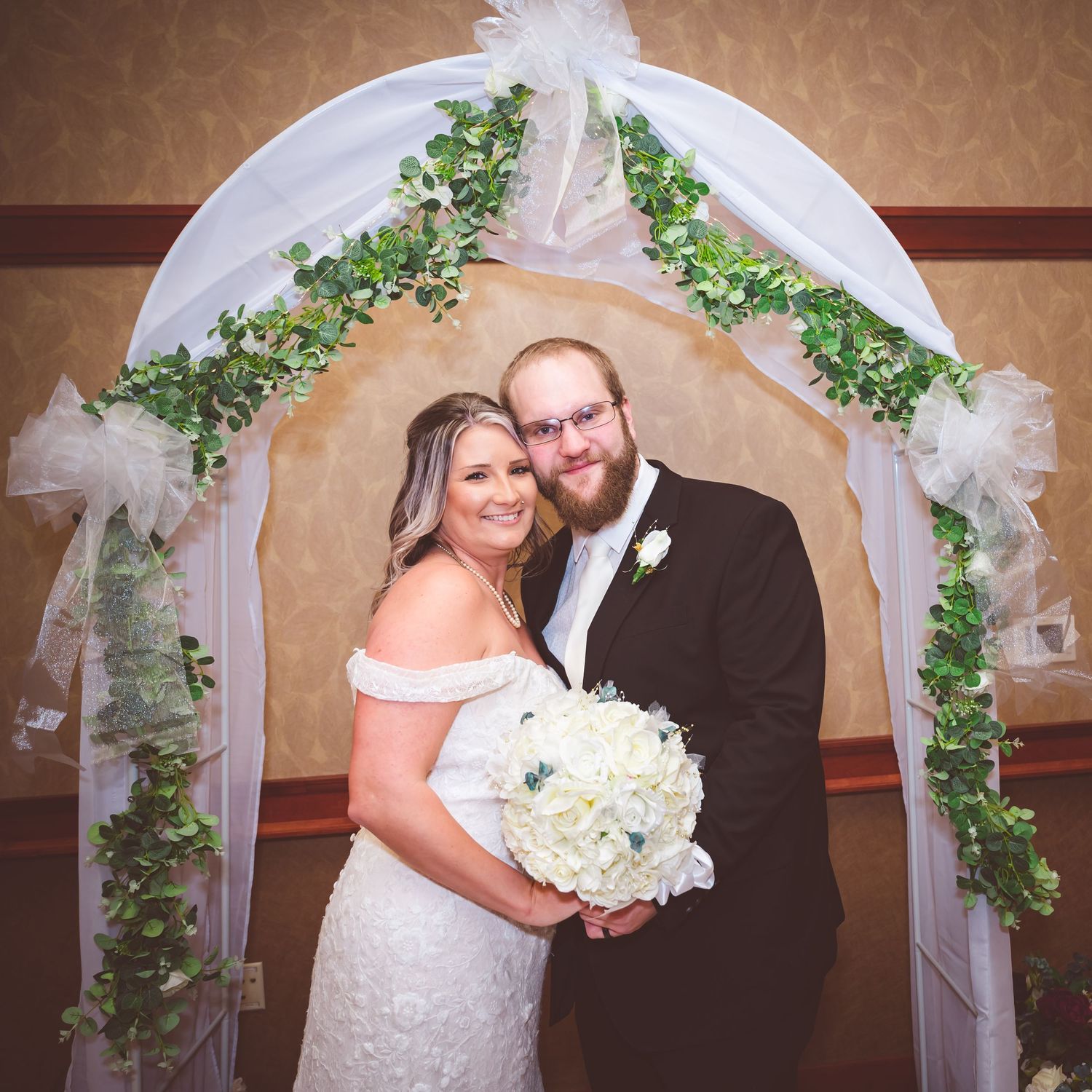 Wedding couple poses under decorative white arch with greenery and floral accents.