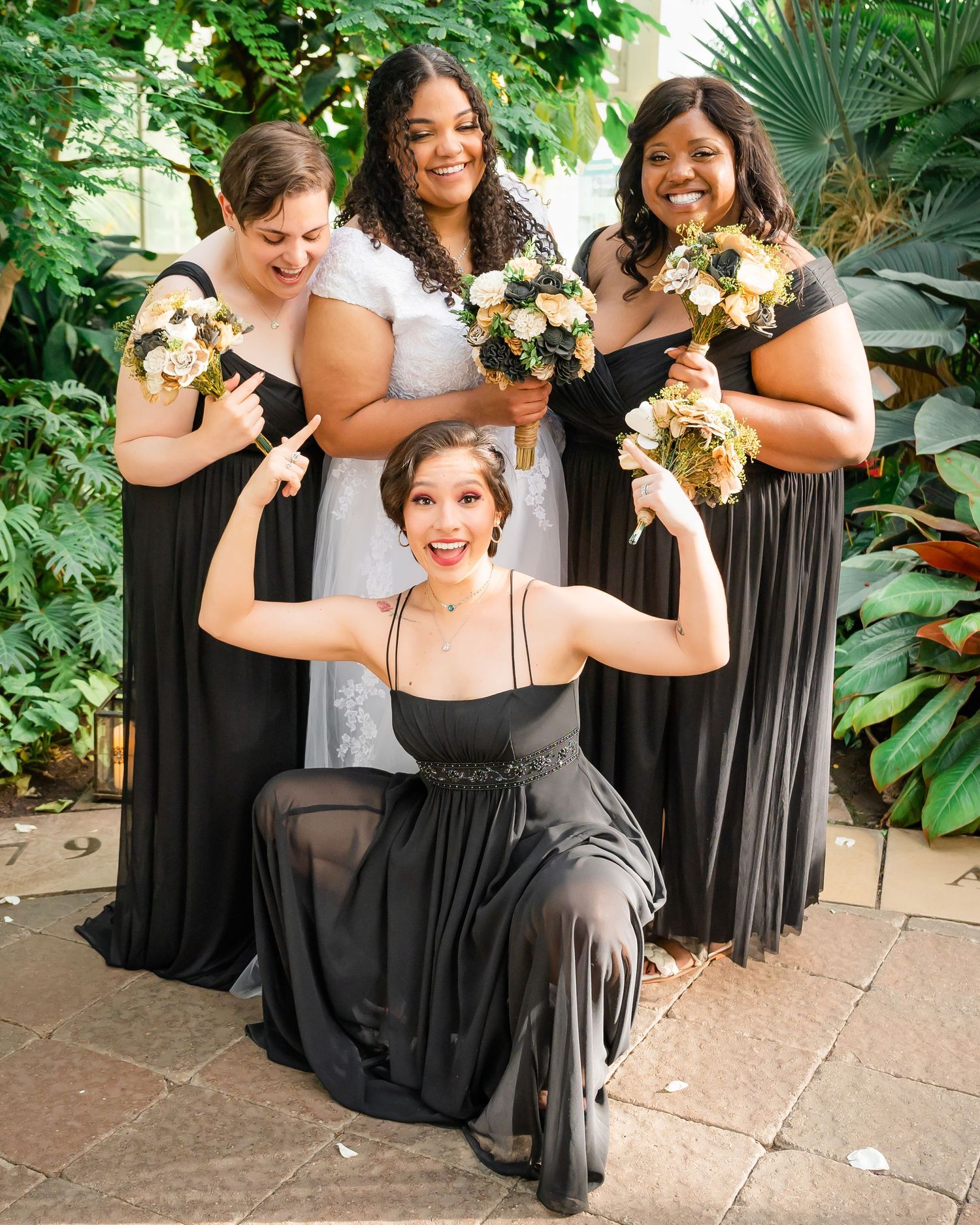 Bridal party in black dresses pose with bouquets against tropical greenery backdrop.