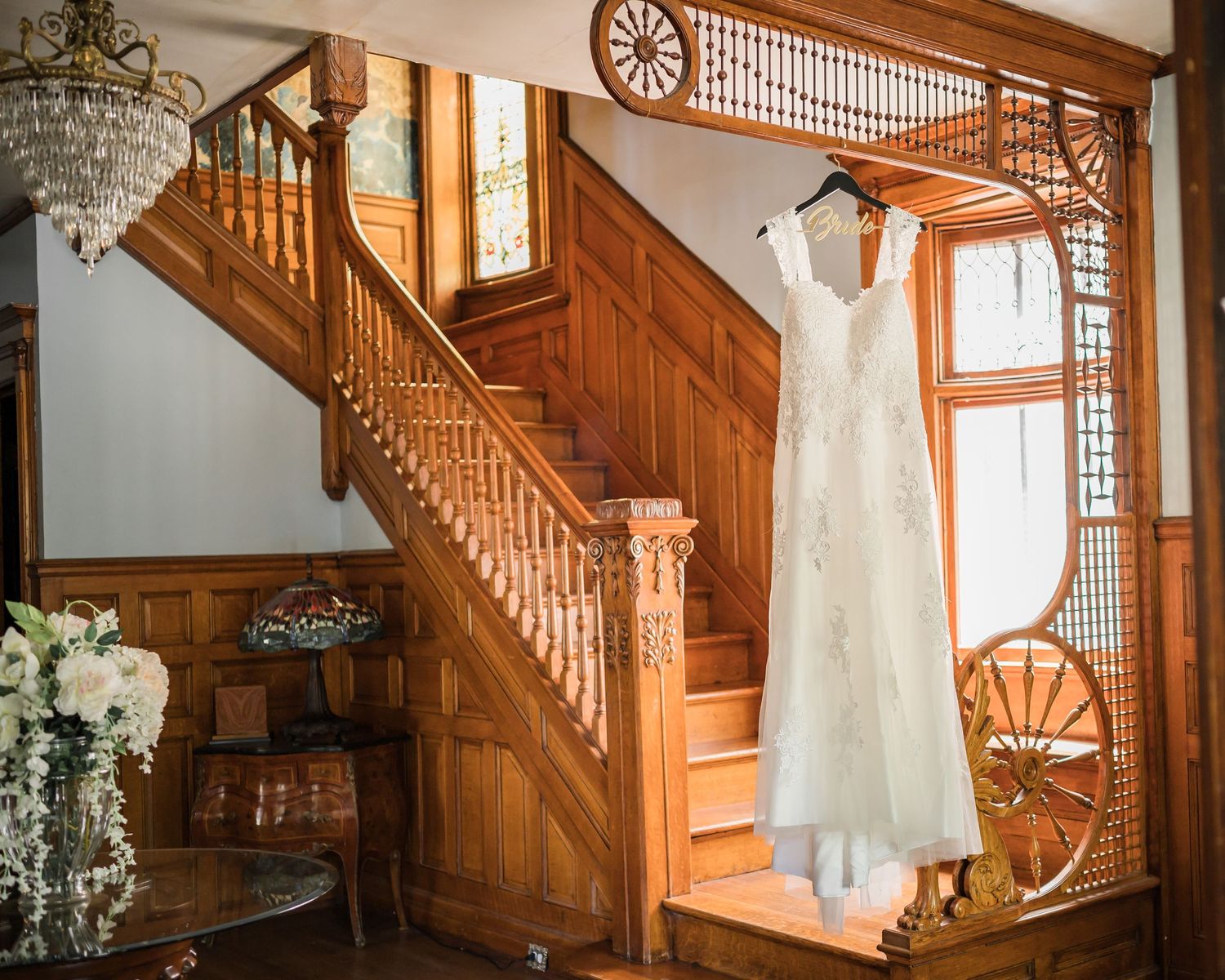 Wedding dress displayed near ornate wooden staircase with crystal chandelier and floral arrangements.