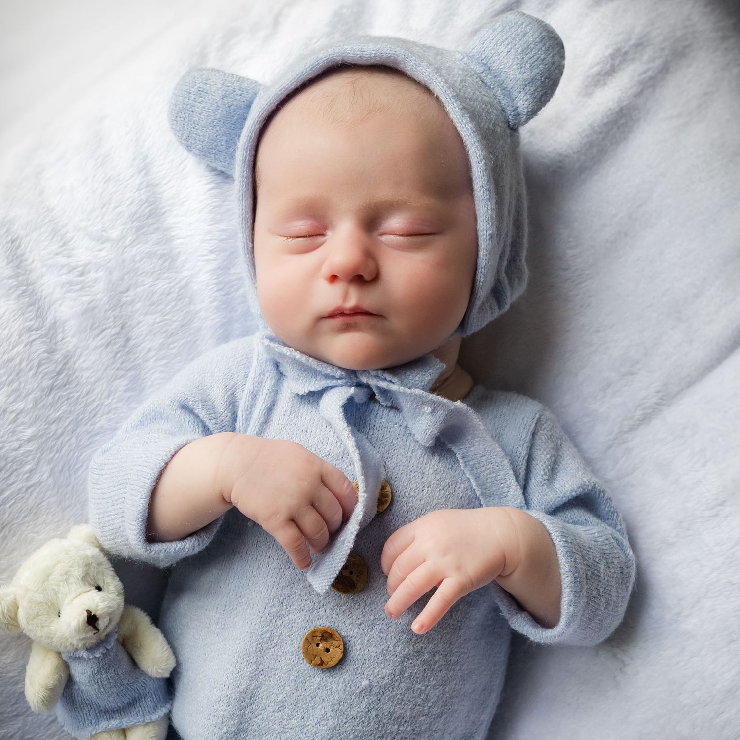 Sleeping baby in soft blue bear outfit lying on white blanket with small plush toy.