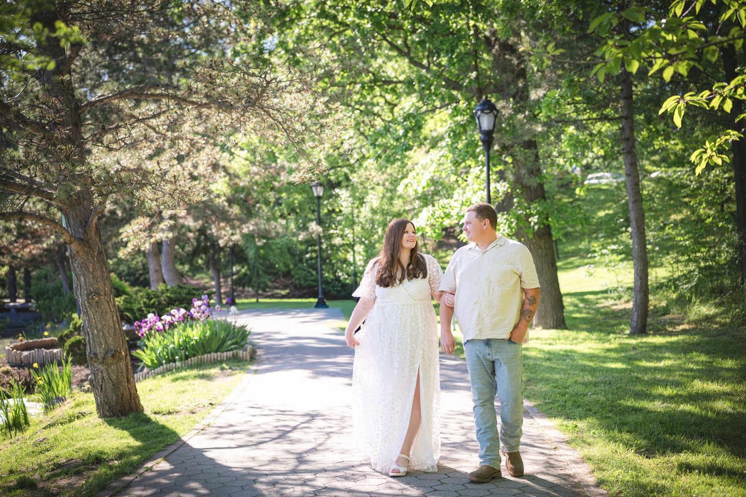 Couple holding hands walking on a tree-lined park path, woman in white maternity dress, man in jeans on sunny day.