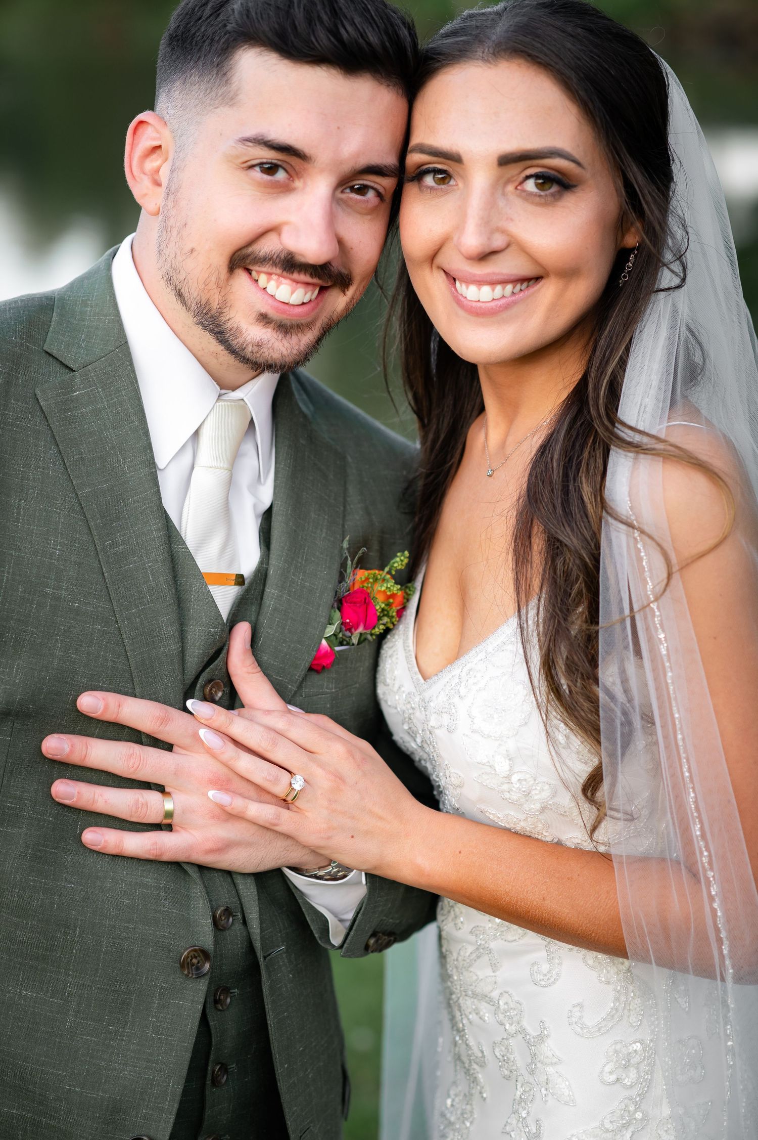 Wedding portrait showing newlyweds in traditional attire with green suit and white dress displaying wedding rings.