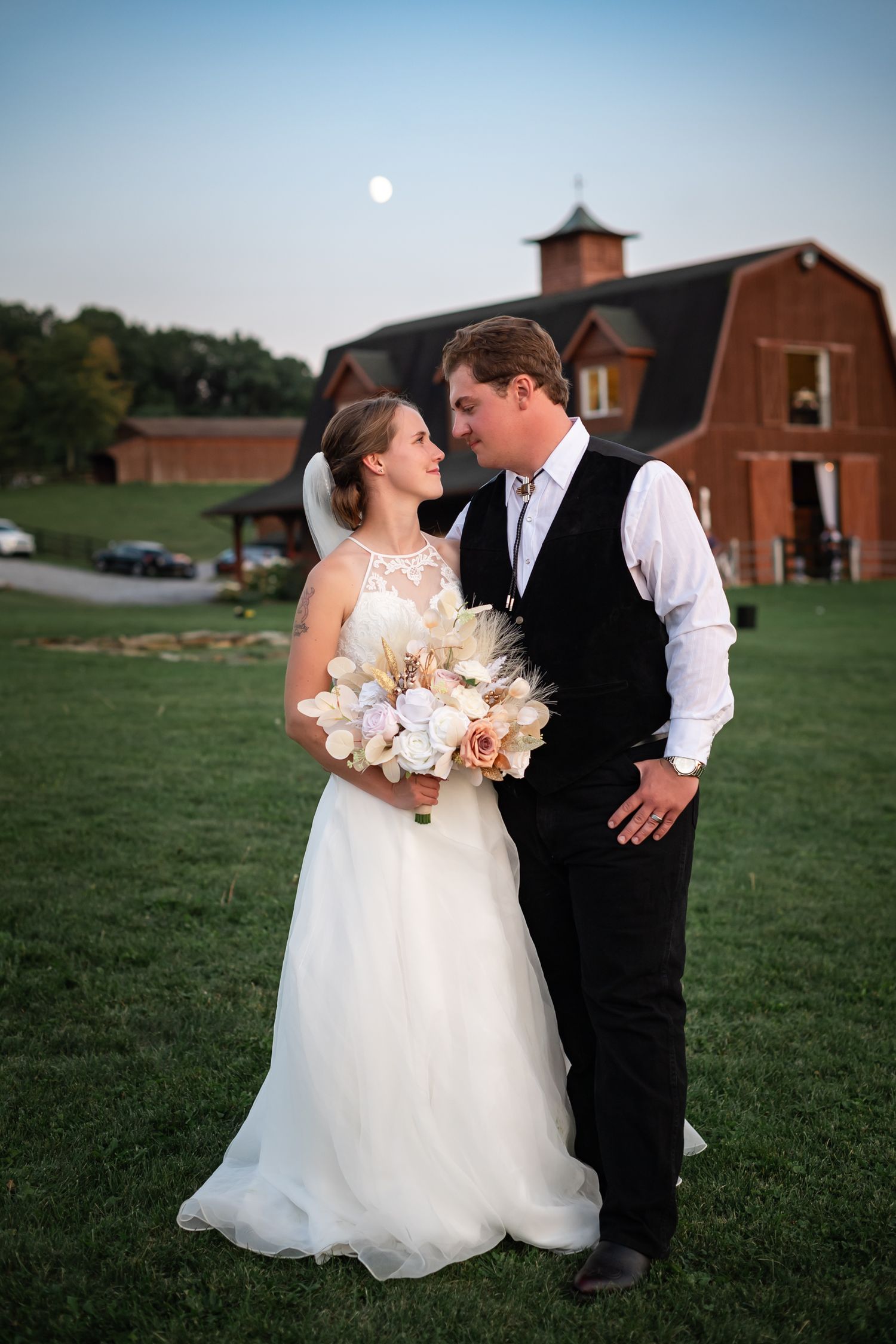 A romantic evening wedding photo outside a rustic red barn with a moon glowing in the dusky blue sky.