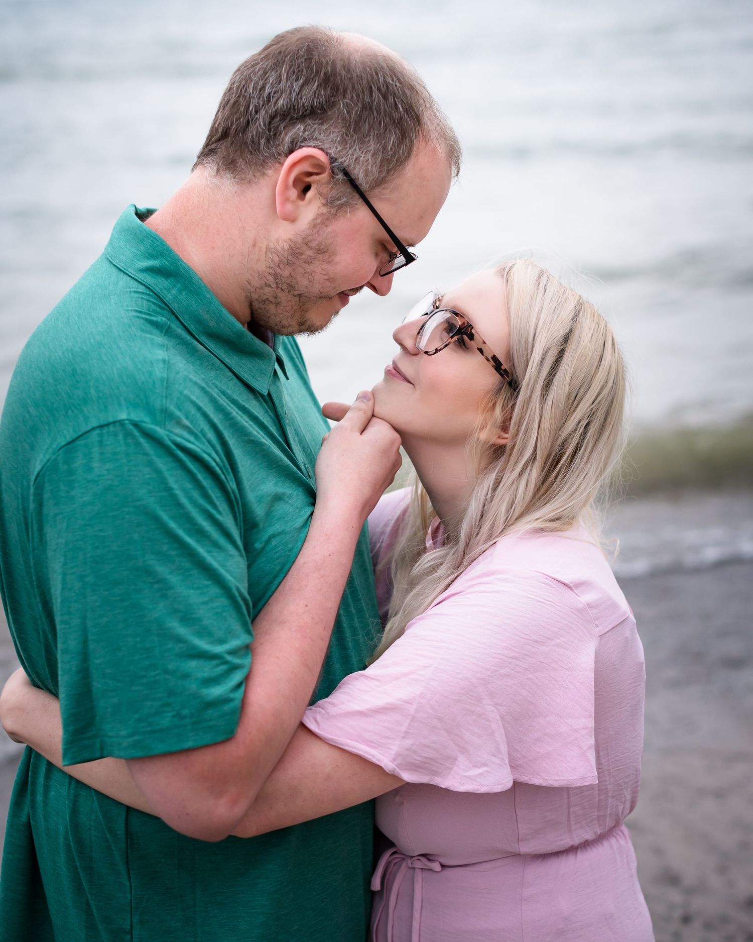 A romantic couple in green and pink shirts share a tender moment on a cloudy day by the water.