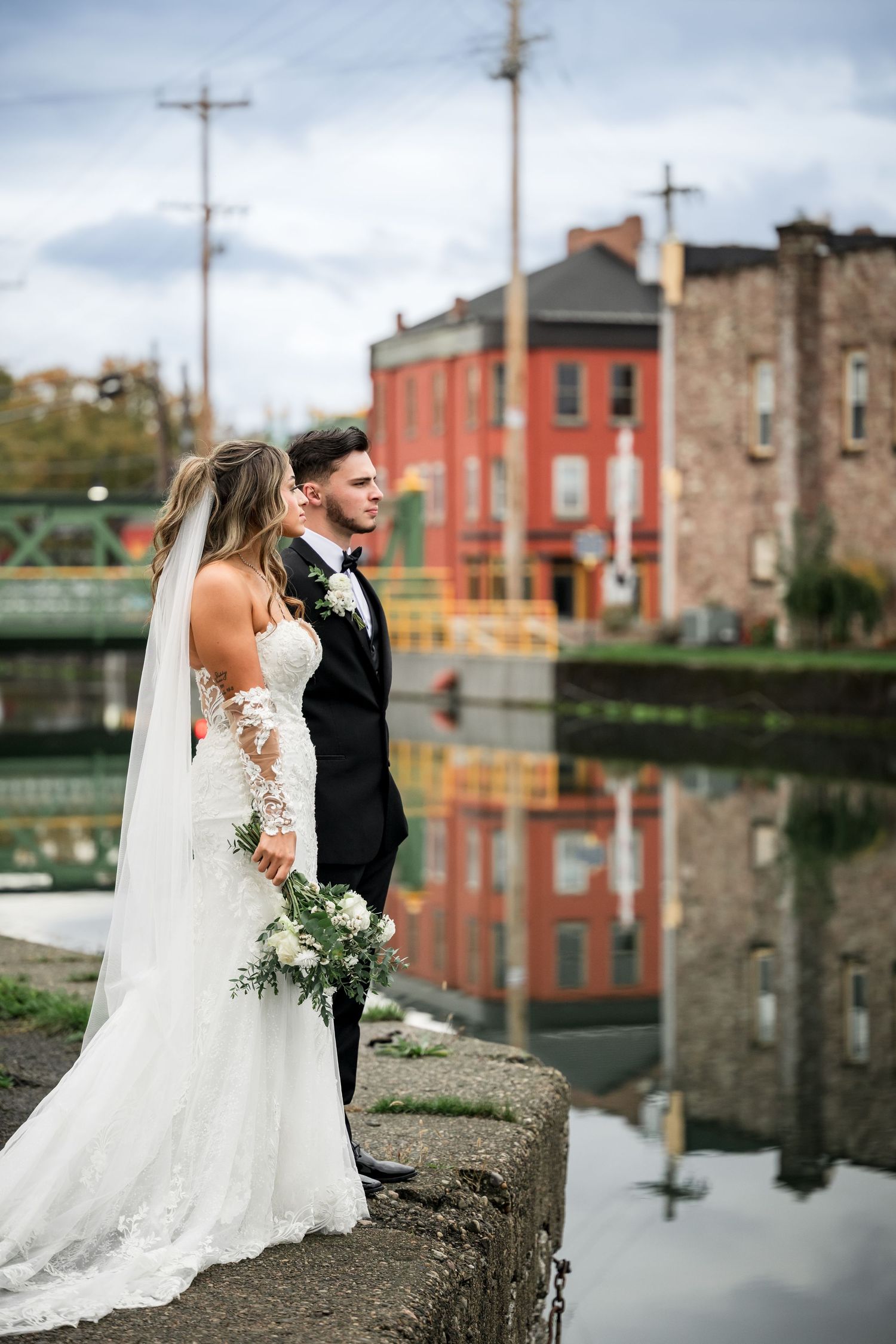A romantic wedding couple stands by a canal with brick buildings and a bridge reflected in the calm water.