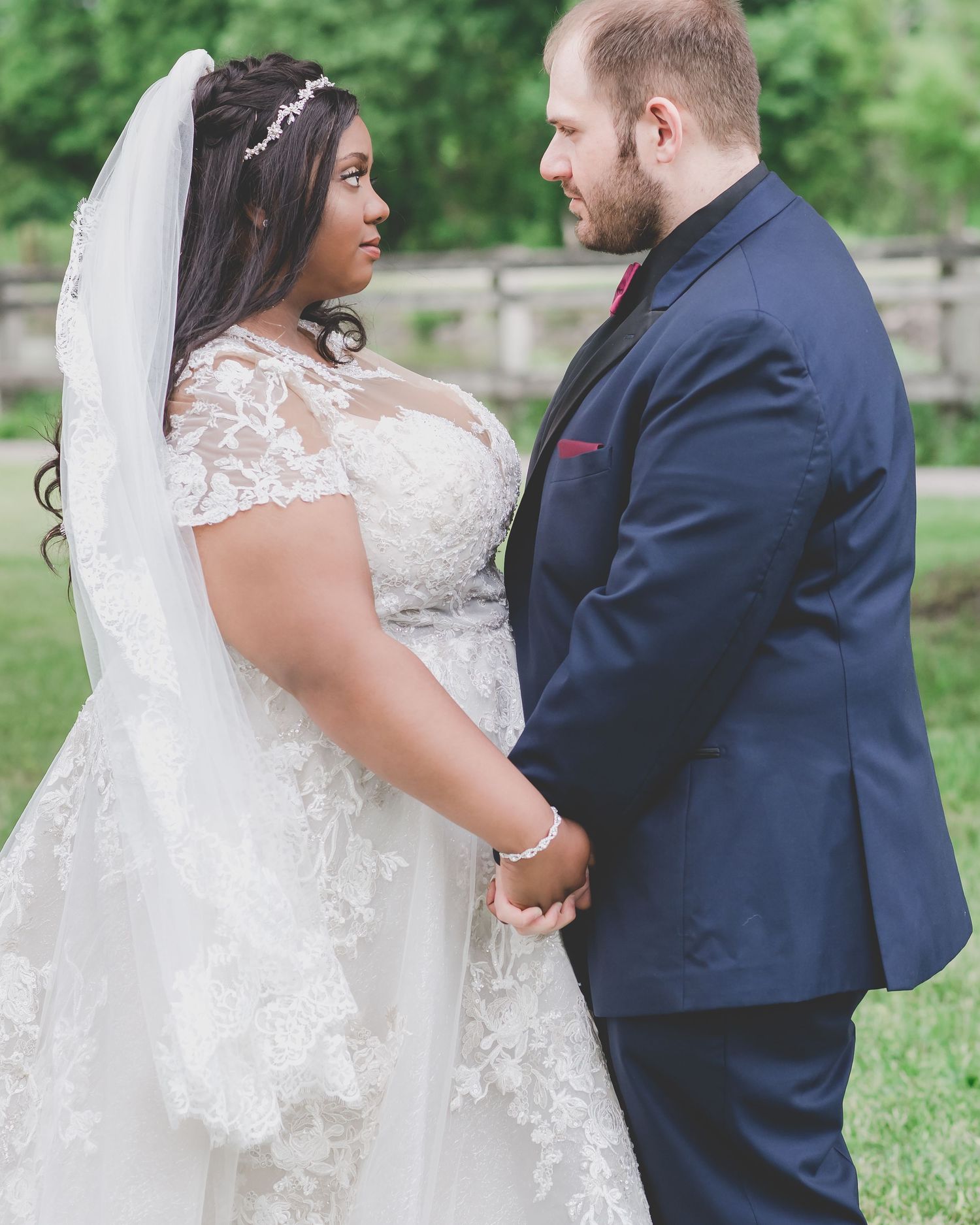 Couple holds hands during wedding ceremony, bride in lace gown and groom in navy suit with red accent.