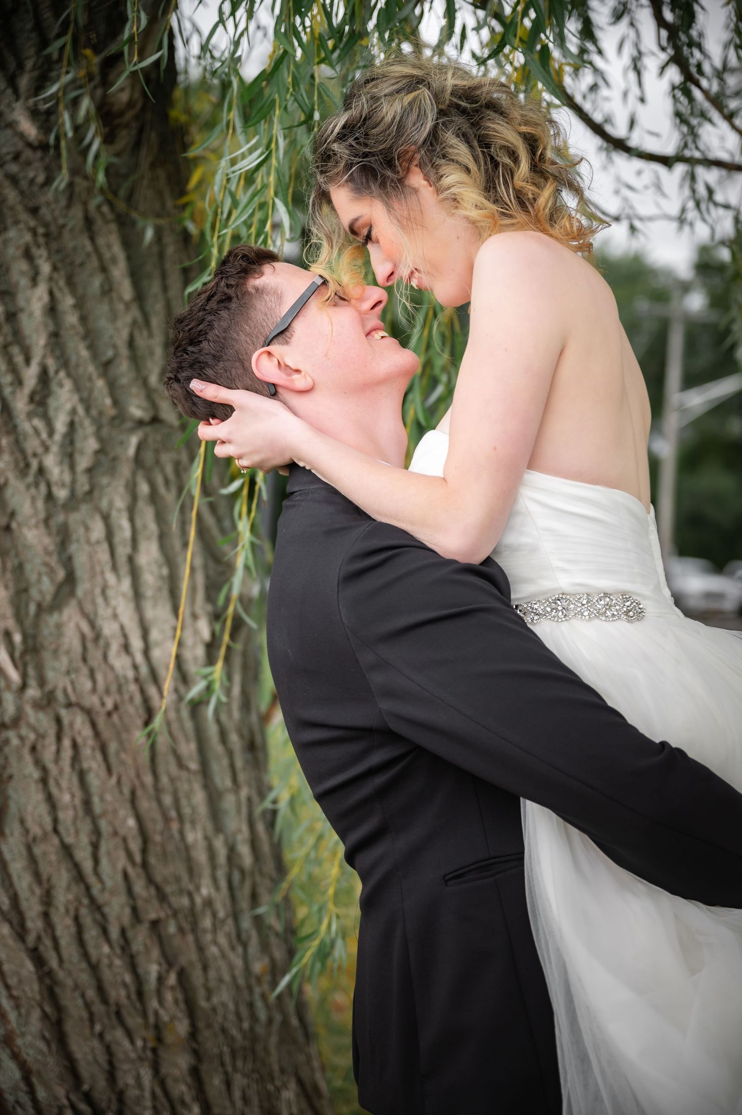 A romantic wedding photo of a couple embracing under willow tree branches during an outdoor ceremony.