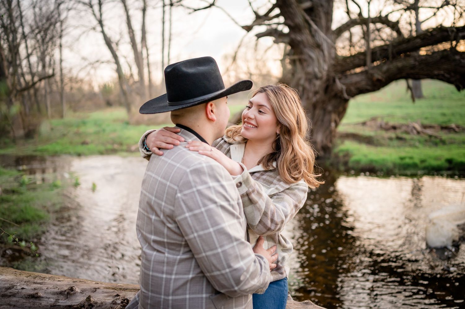 A couple shares an intimate moment outdoors by a reflective stream at sunset while wearing casual country attire.