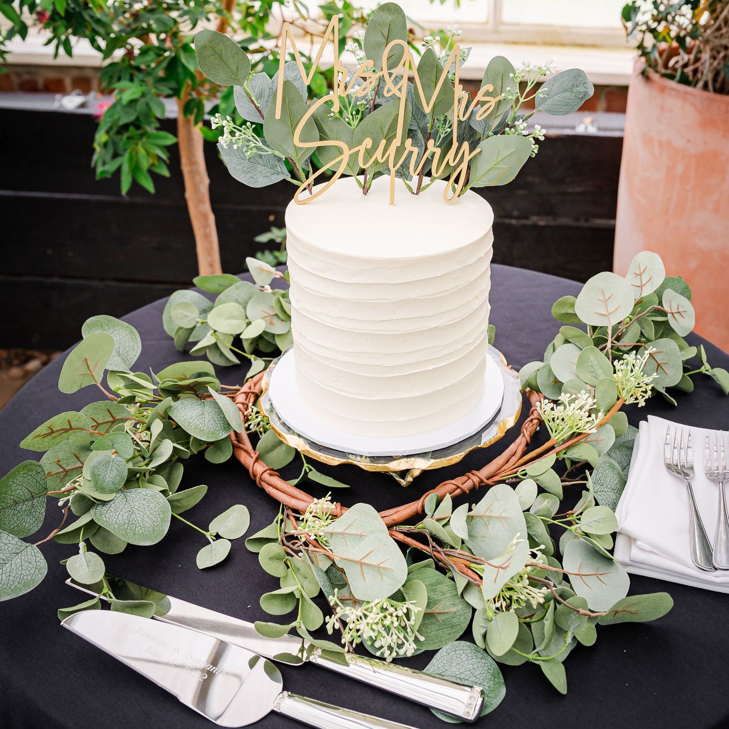 White wedding cake with gold topper decorated with fresh eucalyptus greenery on dark table setting.