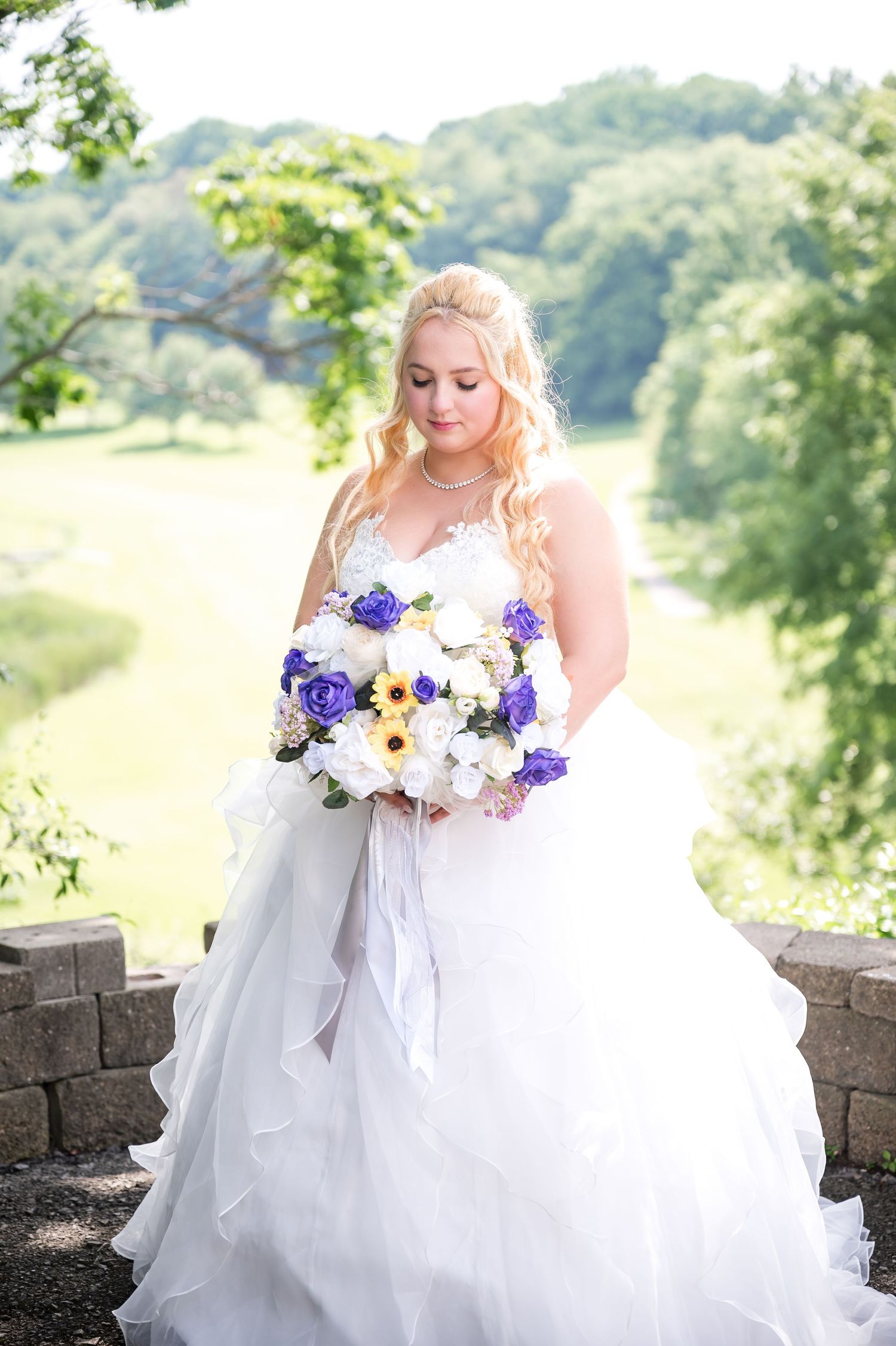 Bride in white gown holding purple and yellow bouquet against scenic hillside backdrop.