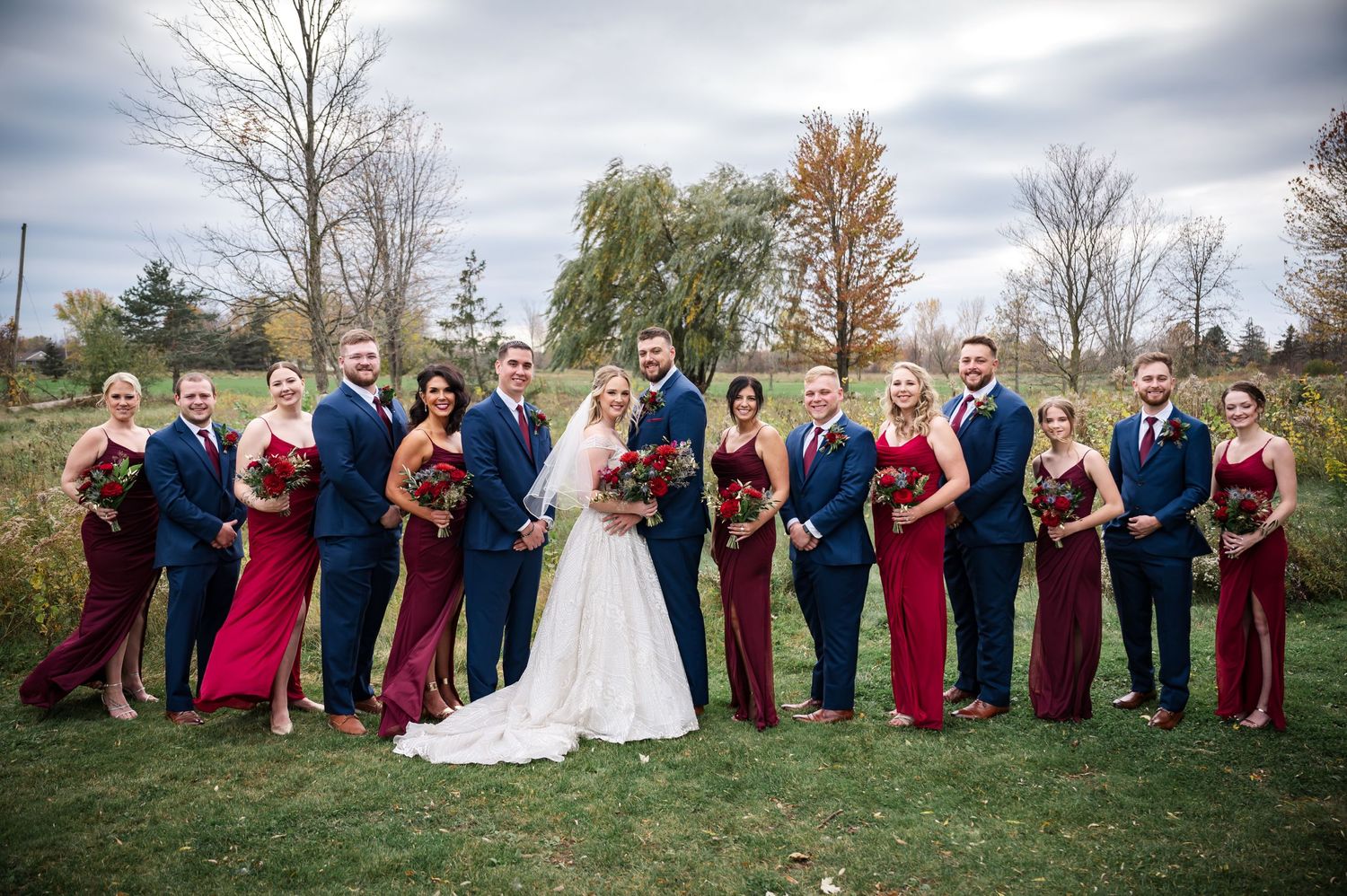 Wedding party poses outdoors in navy blue suits and burgundy dresses against autumn trees and cloudy sky.