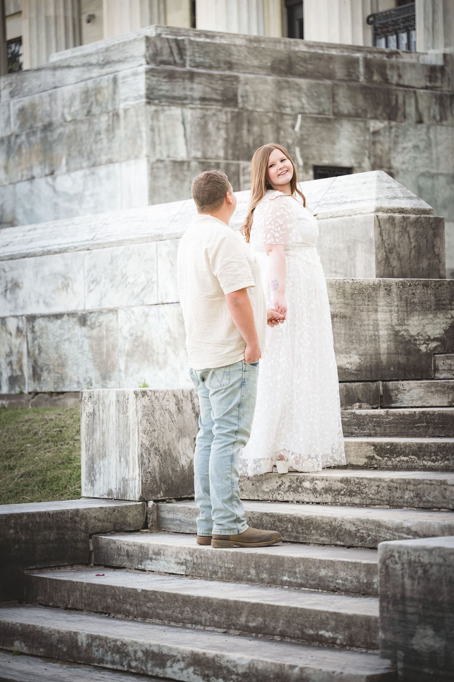 Couple on stone steps, woman in white lace gown smiling at man in casual attire, elegant building backdrop.