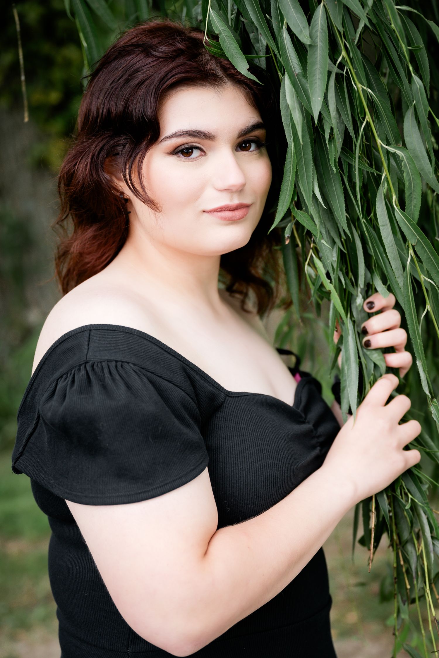 Portrait of someone in a black off-shoulder dress posing elegantly against willow tree branches in a garden setting.
