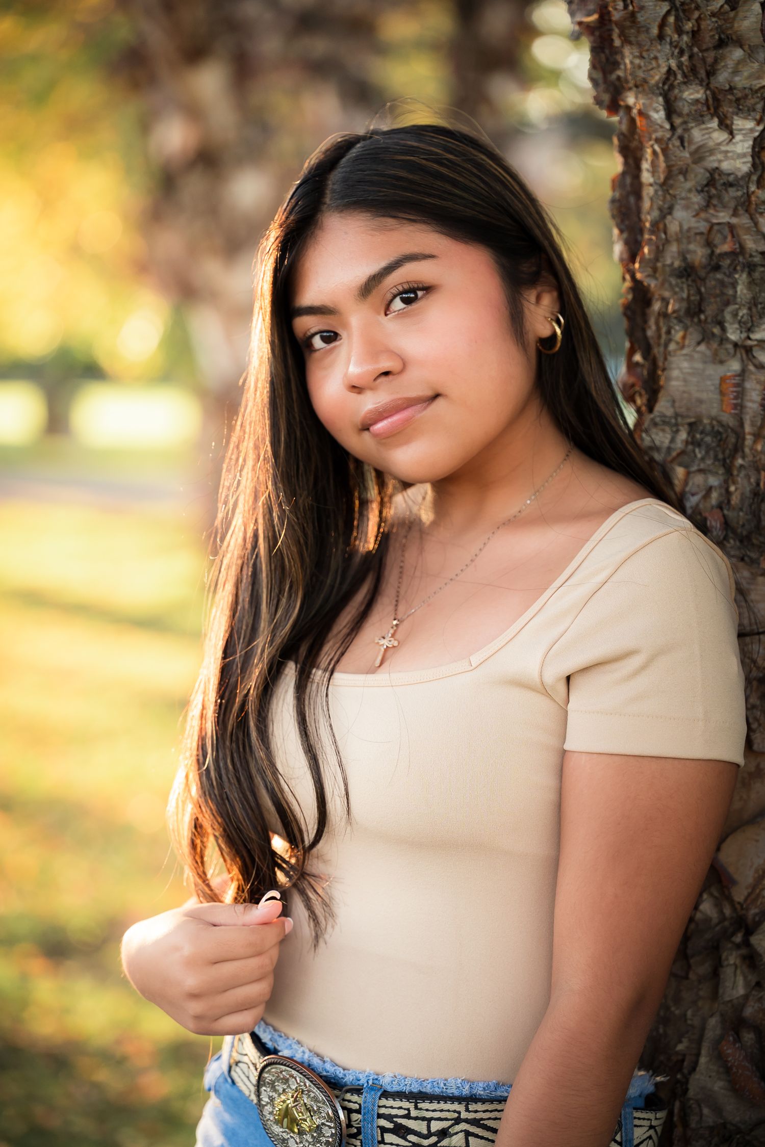 Portrait of someone in a beige top leaning against a tree trunk with warm autumn lighting in the background.