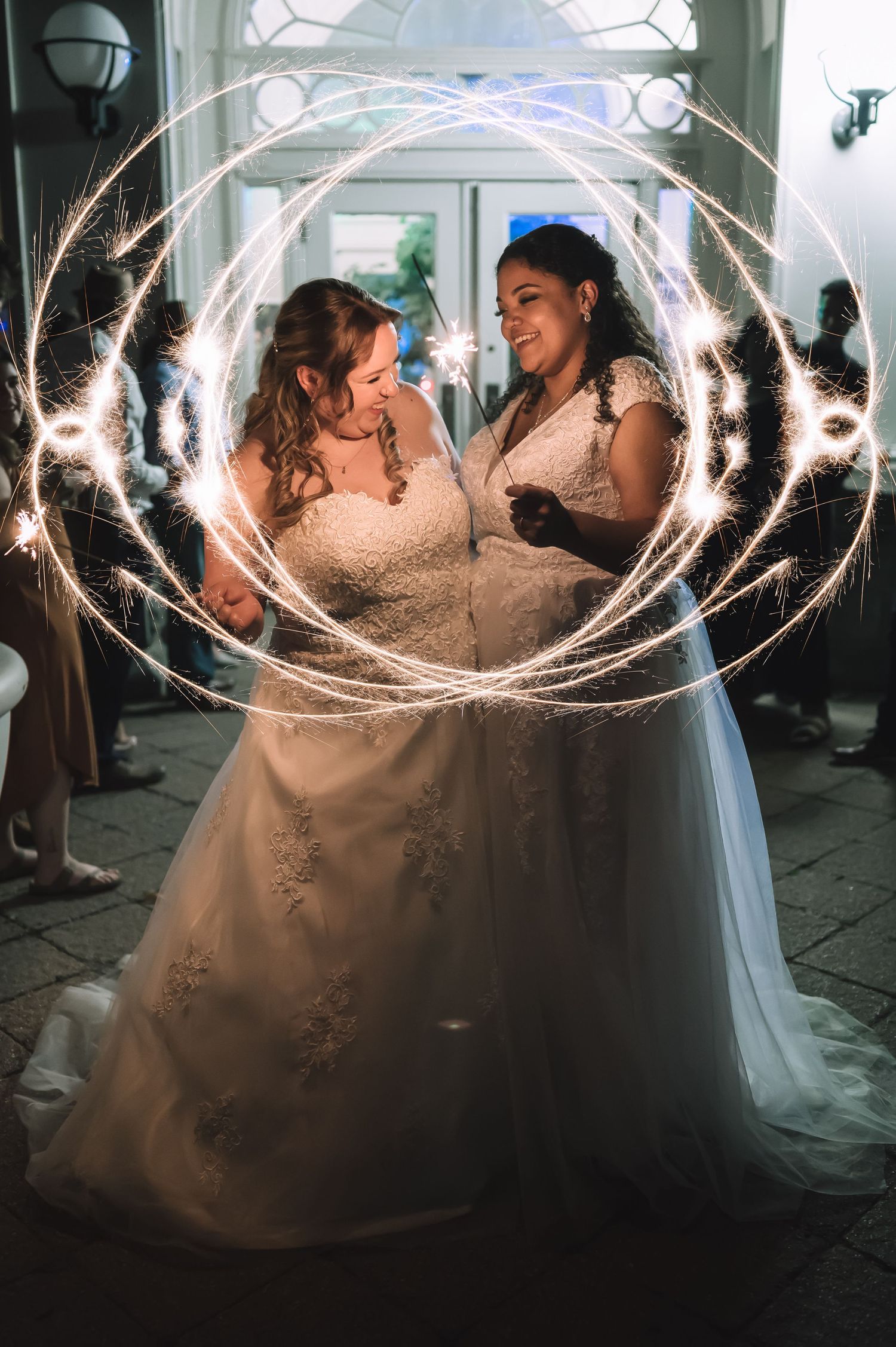 Wedding couple in white dresses surrounded by sparkler light effects creating circular patterns.
