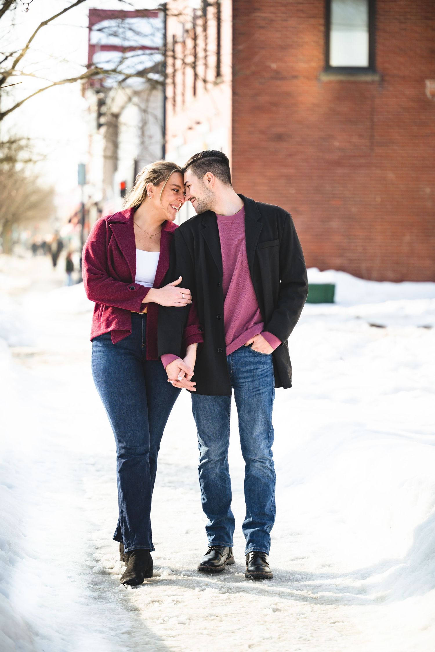 Smiling engaged couple walking hand in hand on snowy urban sidewalk.