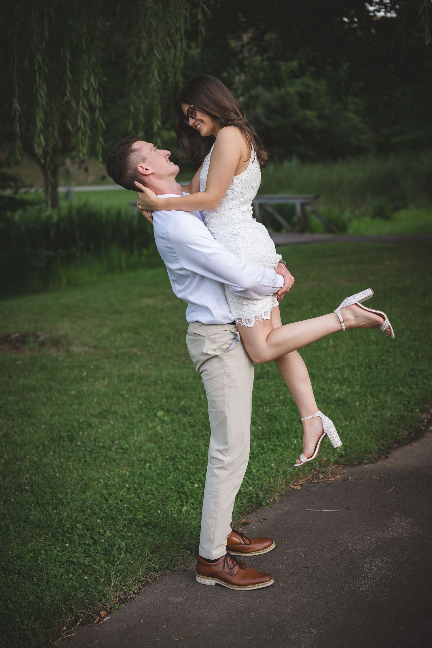 A man lifts a woman in a white dress in a lush green park, sharing a joyful romantic moment outdoors.