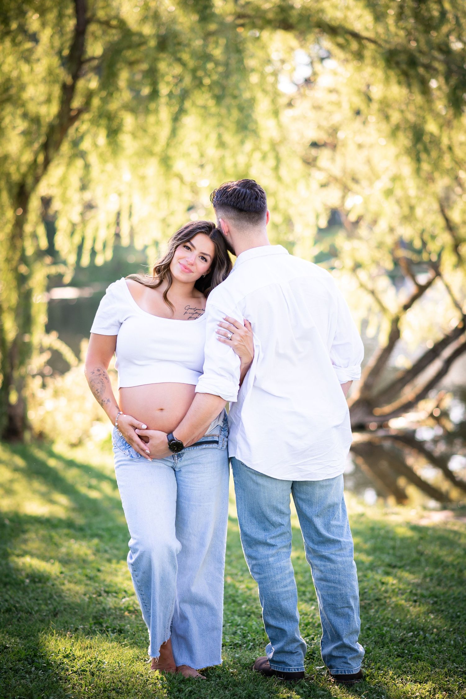 Pregnant couple in white tops and jeans pose outdoors under willow tree, woman cradling baby bump.