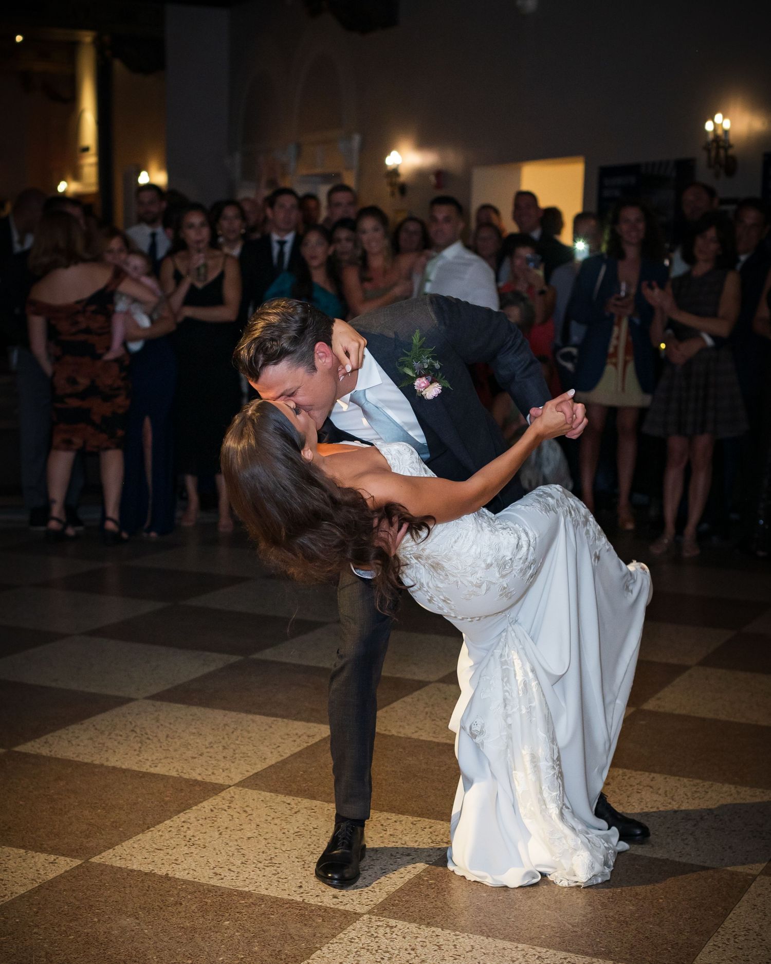 Couple performs romantic dip during their first dance at wedding reception while guests watch from background on dance floor.