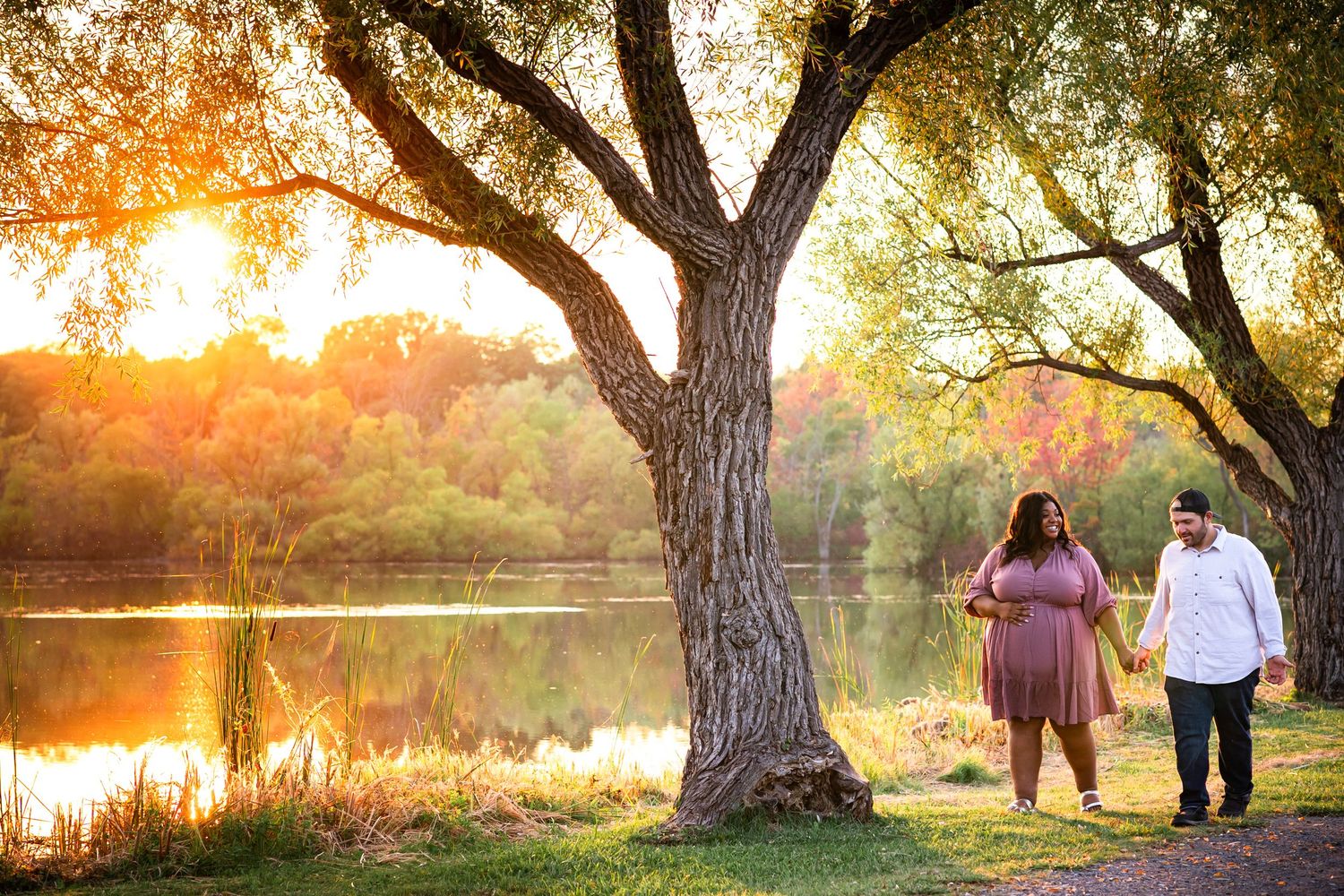 Silhouetted figures hold hands by a large tree during golden hour near a reflective lake.
