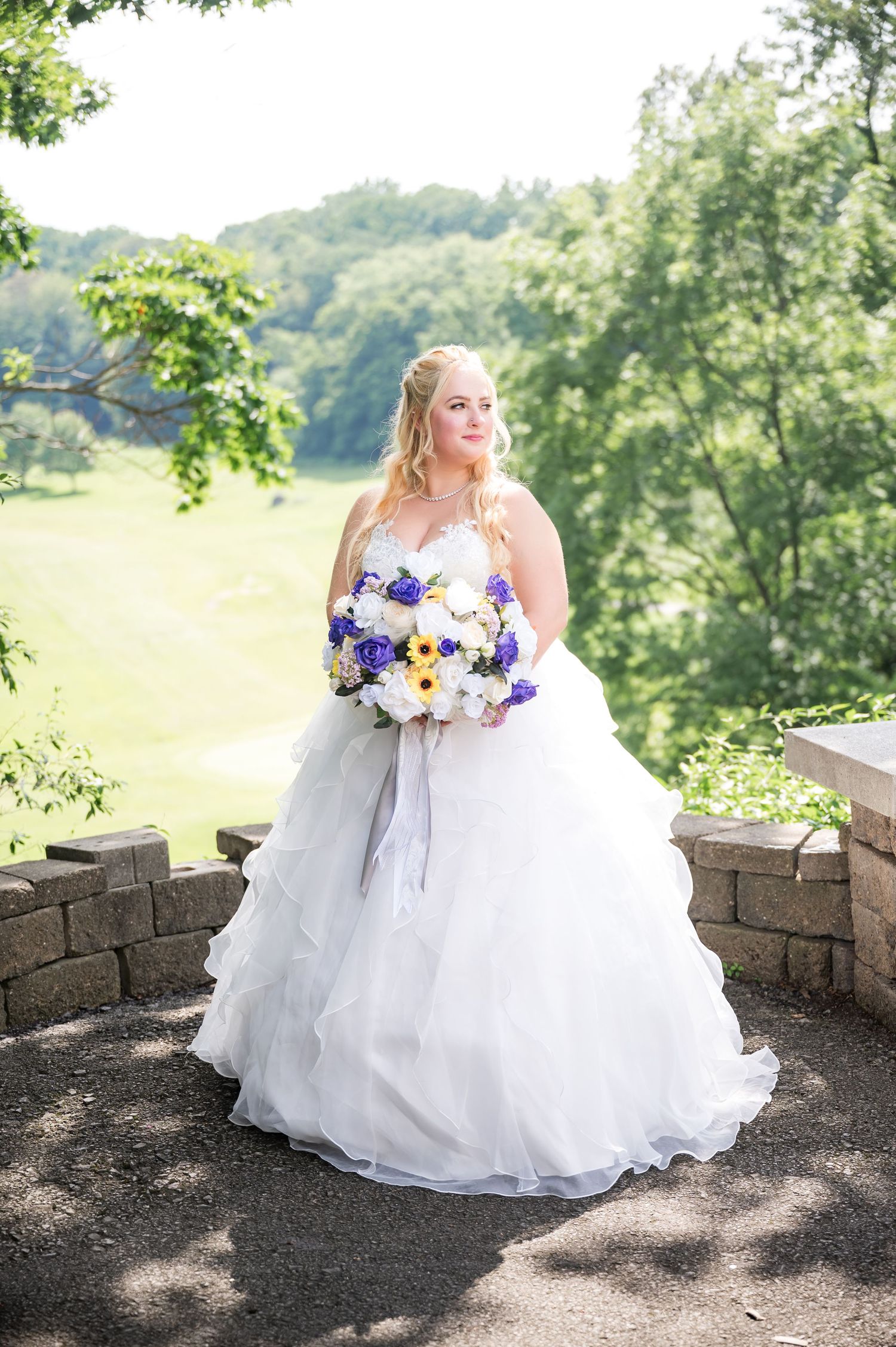 A bride in a white ballgown holds a purple and yellow bouquet while standing on a scenic overlook.