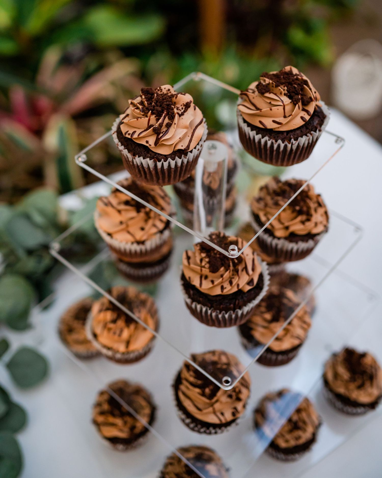 Chocolate cupcakes with brown frosting arranged on a tiered display stand with greenery in background.