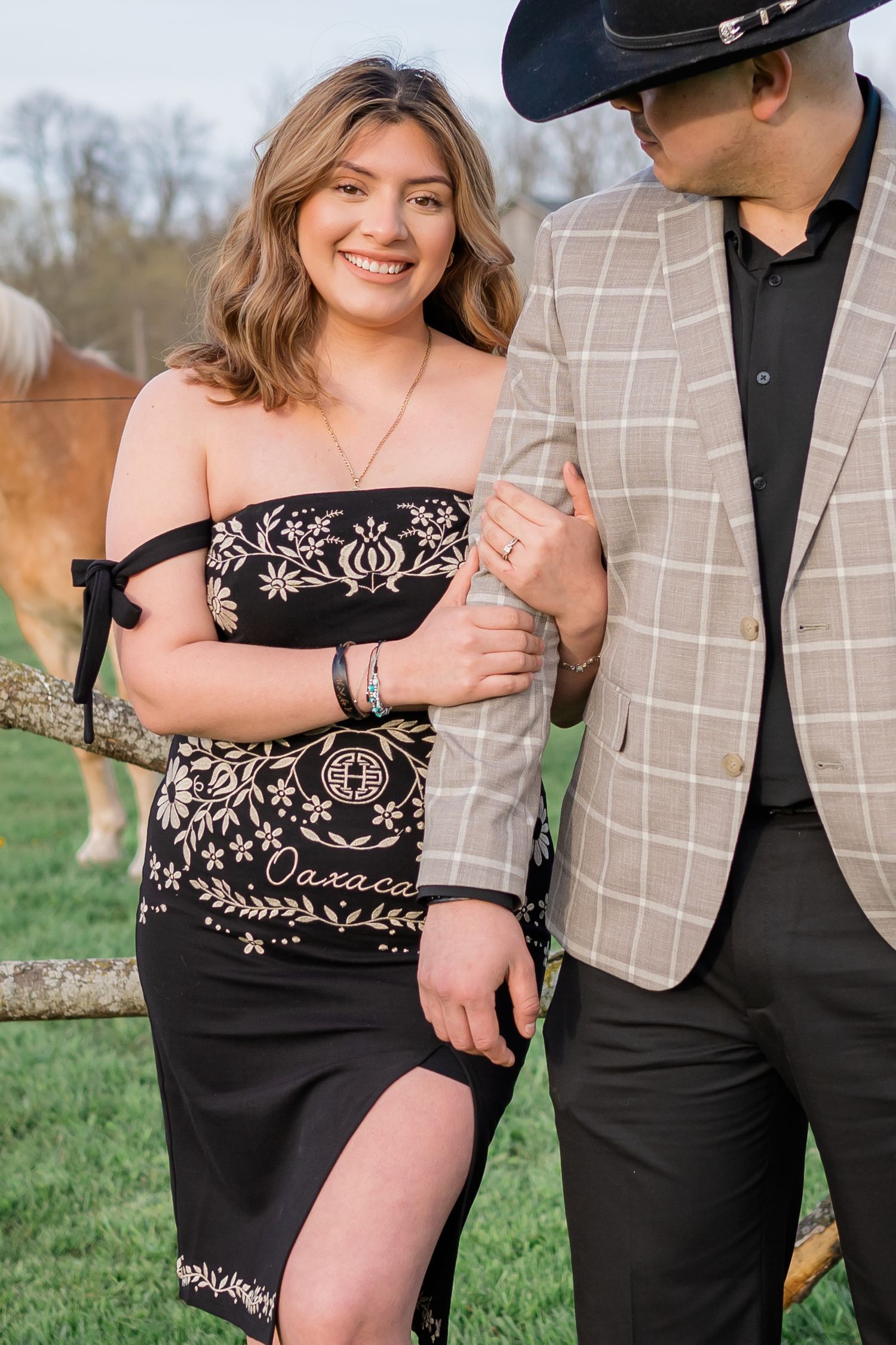 Romantic couple in country western attire pose together by rustic fence on grassy field at sunset.