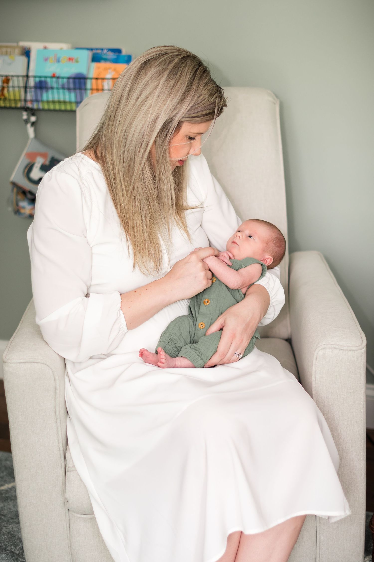 A person in a white dress sits in a light gray armchair feeding a tiny newborn baby wearing sage green clothes.