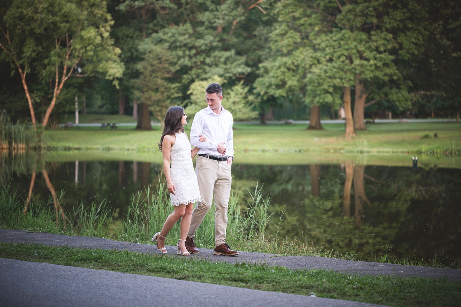 Couple walking hand in hand along a scenic park path beside a tranquil pond during engagement photos.
