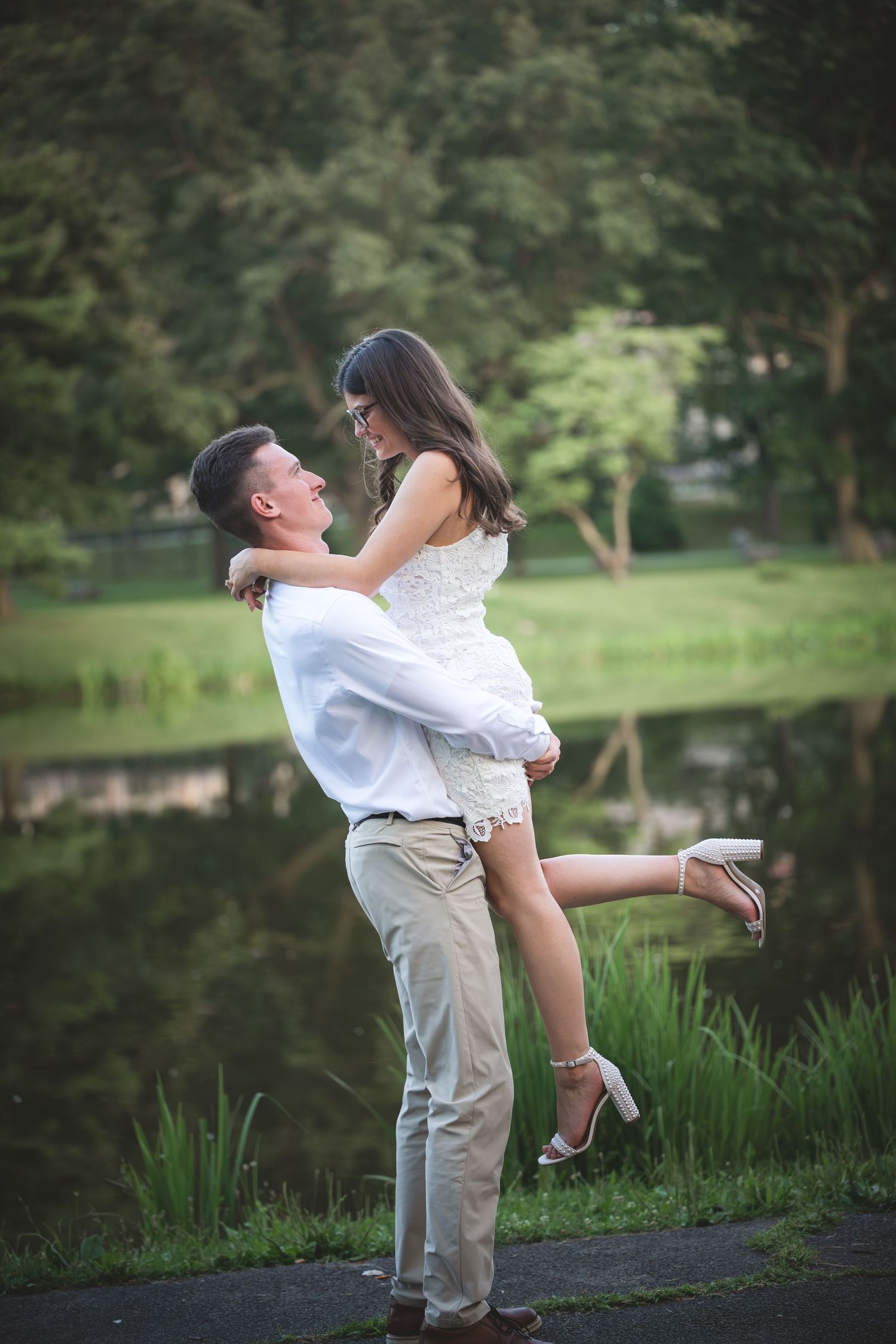 Man lifting woman in white dress by a serene pond in a lush green park, romantic couple portrait.