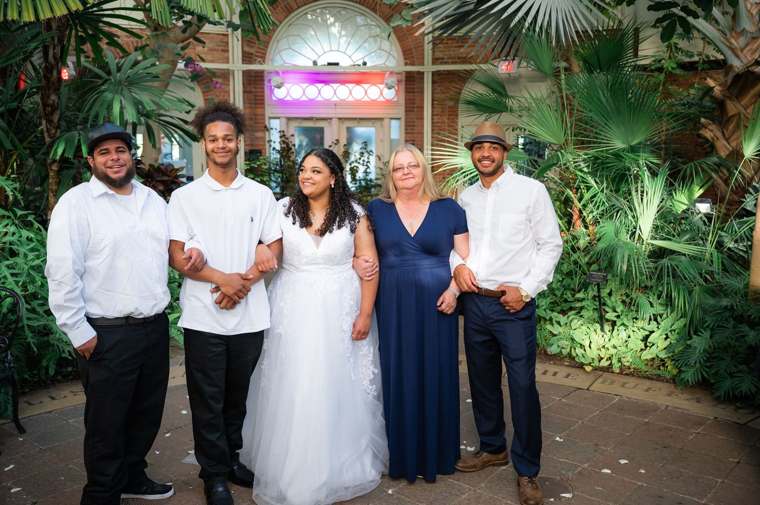 Wedding party members in formal attire pose together against a garden setting with purple lighting.