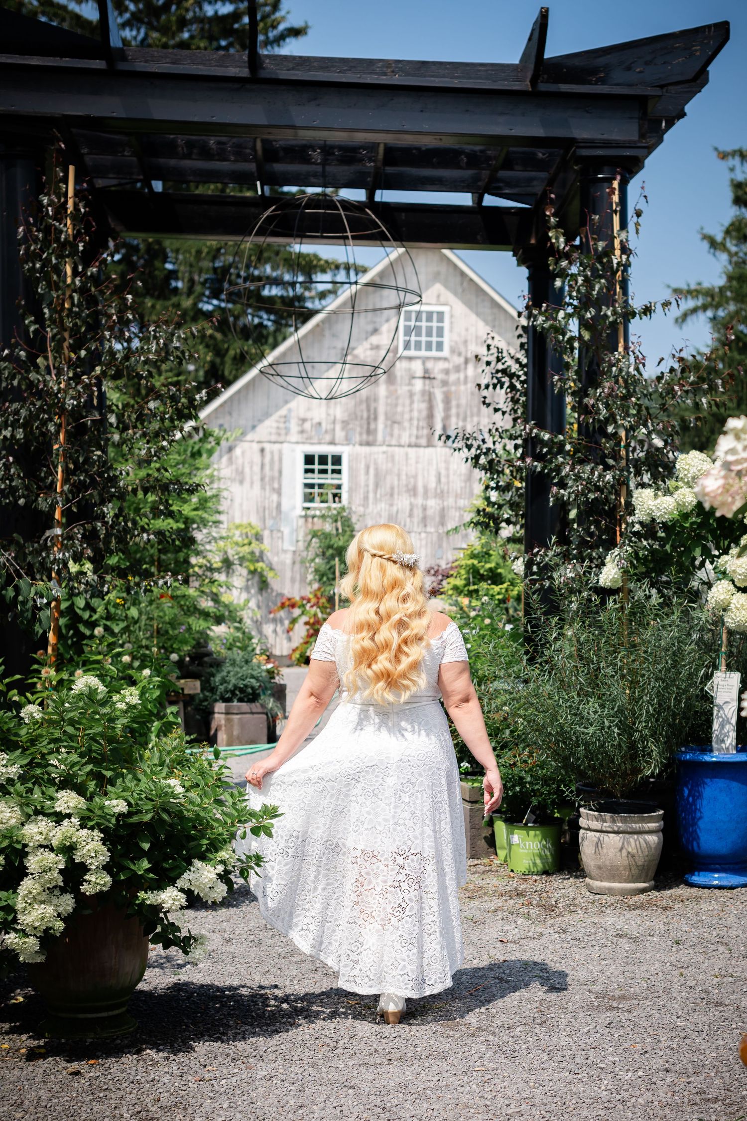 A rustic garden path leads through a wooden arbor towards a weathered barn-style building.