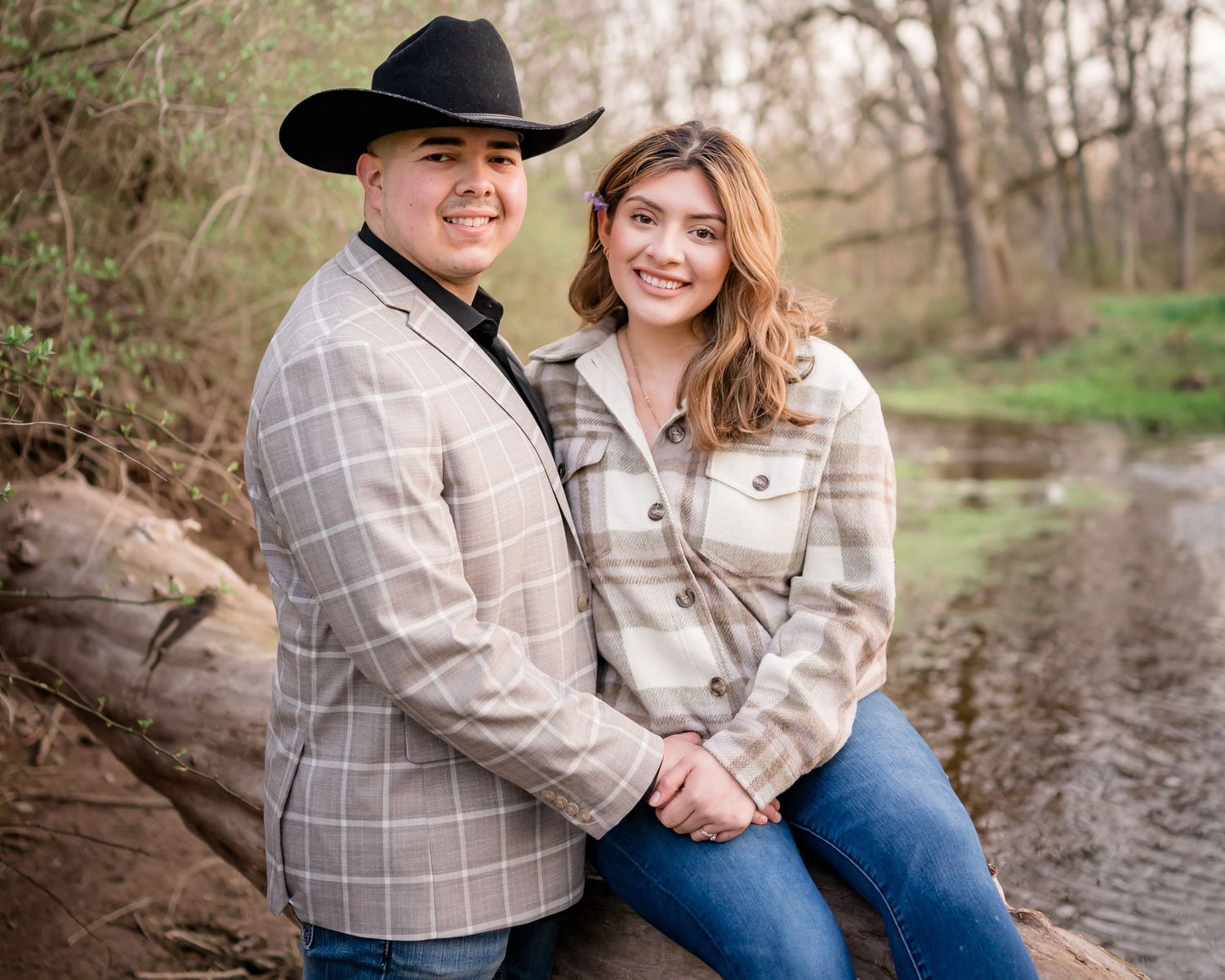 Couple in matching plaid outfits pose together by stream in rural woodland setting.