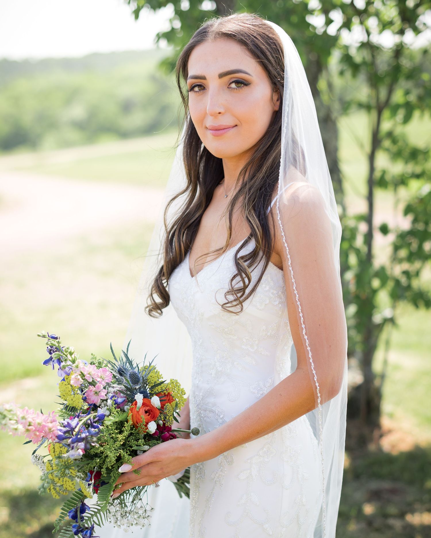 A bride in a white wedding gown and veil holds a vibrant wildflower bouquet in an outdoor garden setting.