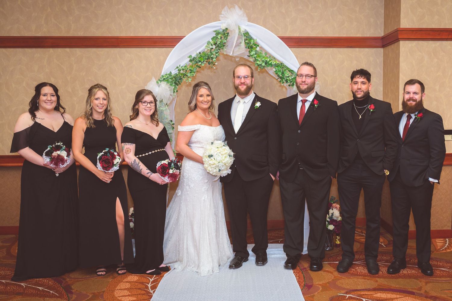 Wedding party poses under a floral arch with bridesmaids in black dresses and groomsmen in dark suits.