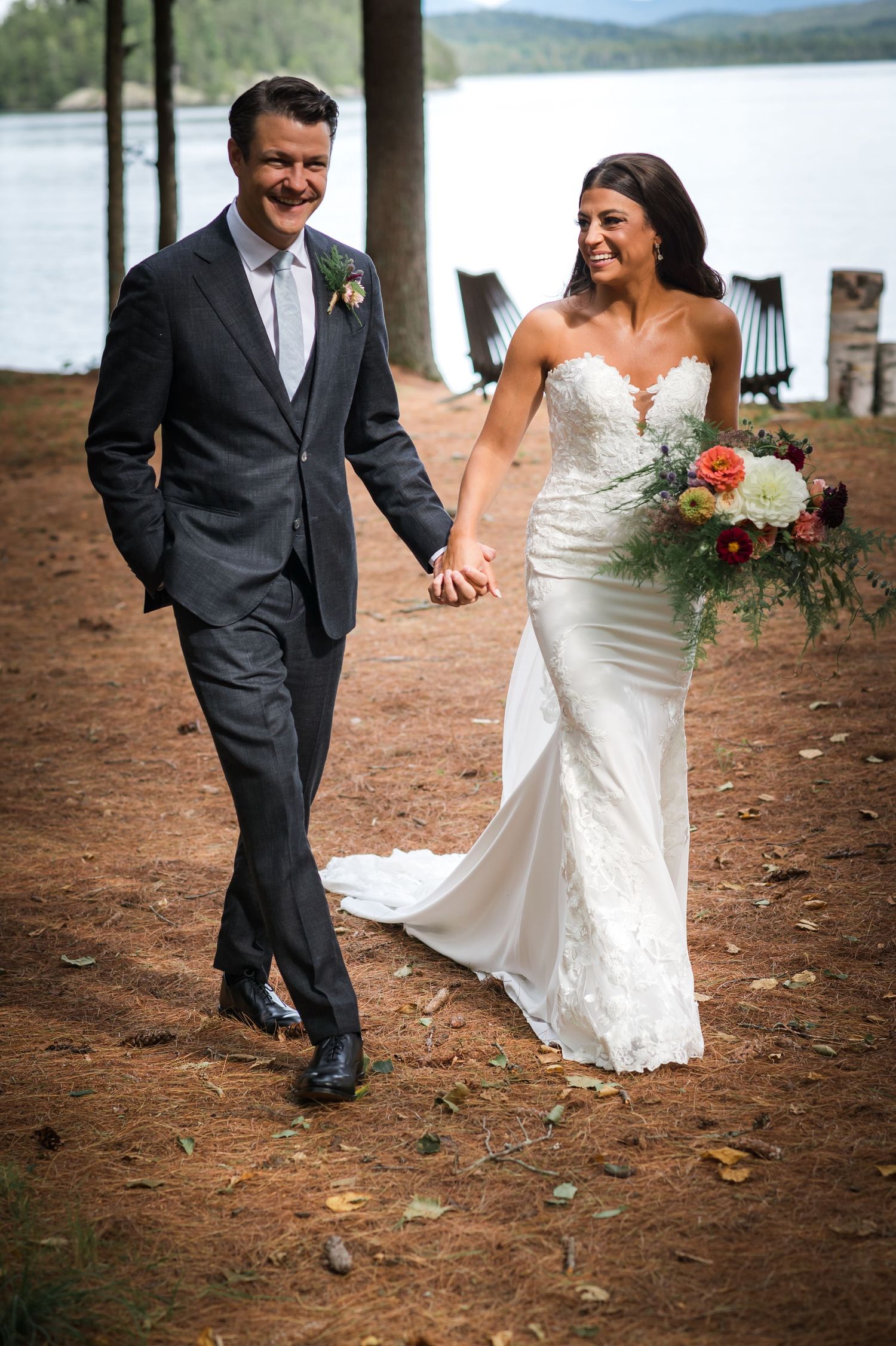 A newlywed couple walks hand in hand along a lakeside trail.