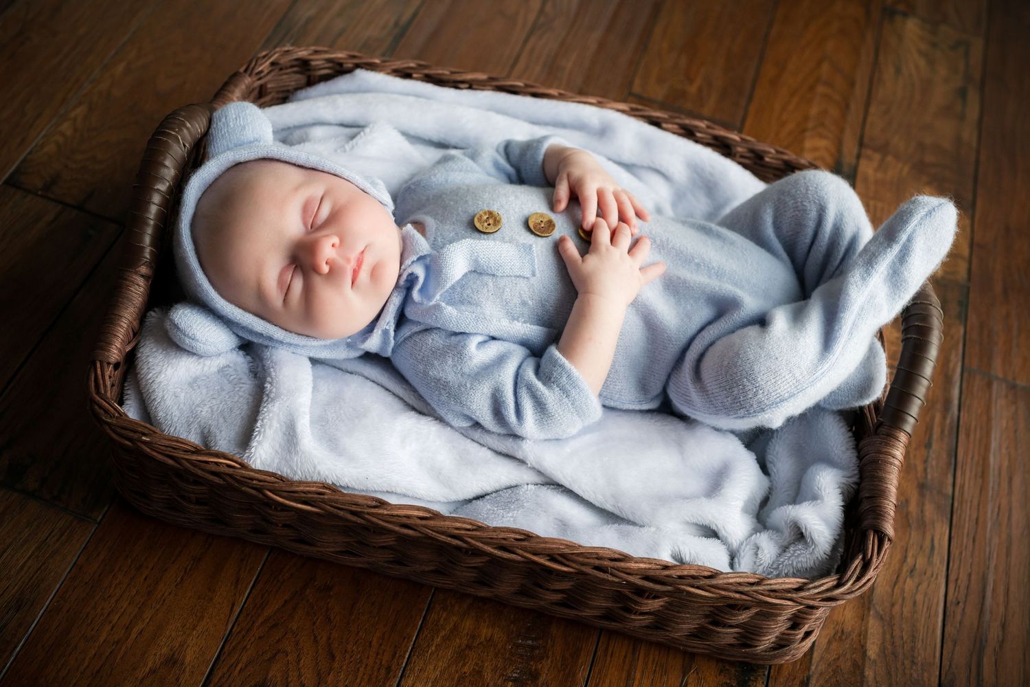 A newborn baby wearing a light blue onesie sleeps peacefully in a woven wicker basket.