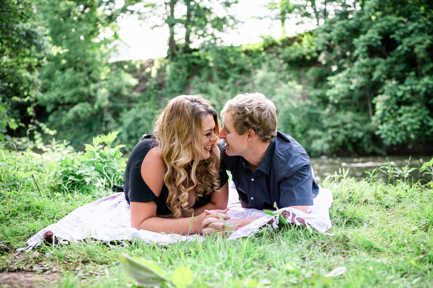 A couple lies on a white blanket in a lush green garden setting while sharing an intimate moment together.