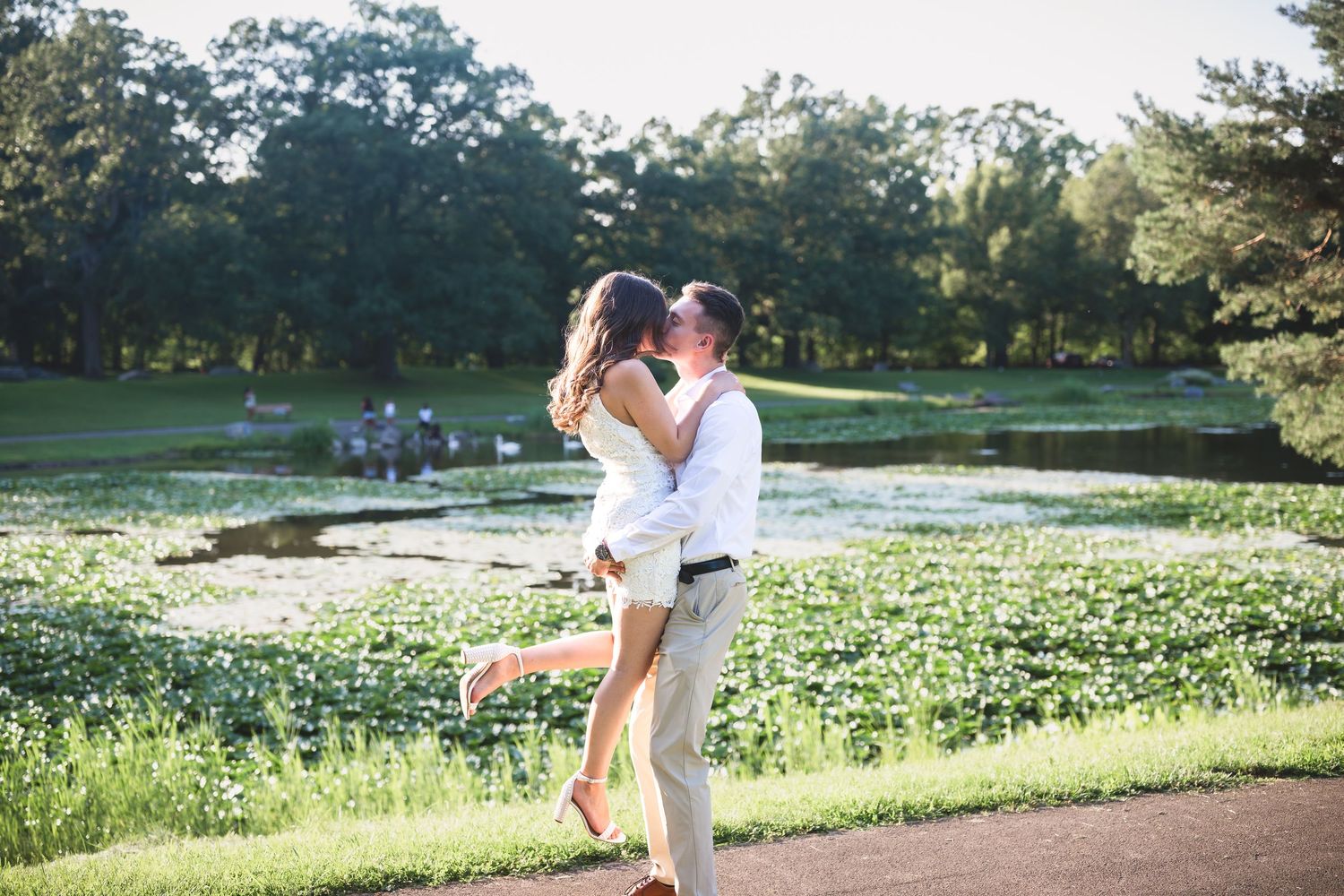 Couple sharing a romantic kiss by a serene pond surrounded by lush greenery on a sunny day.