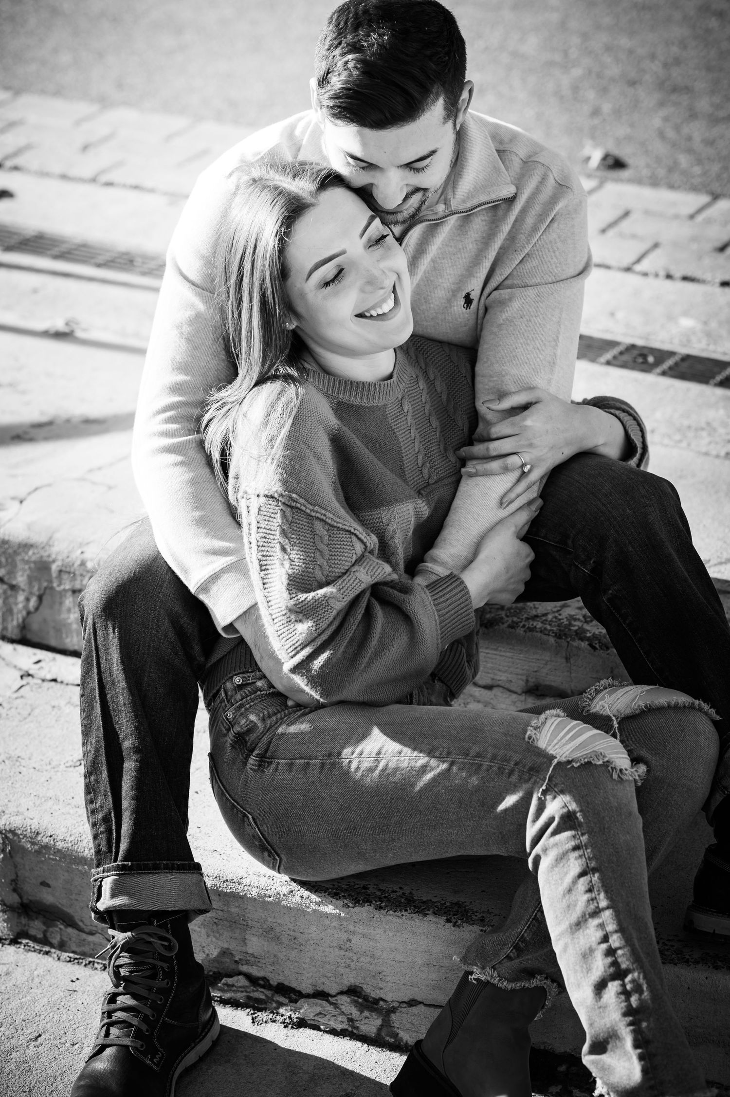 A romantic black and white portrait of a couple embracing on concrete steps, showing tender affection and joy.