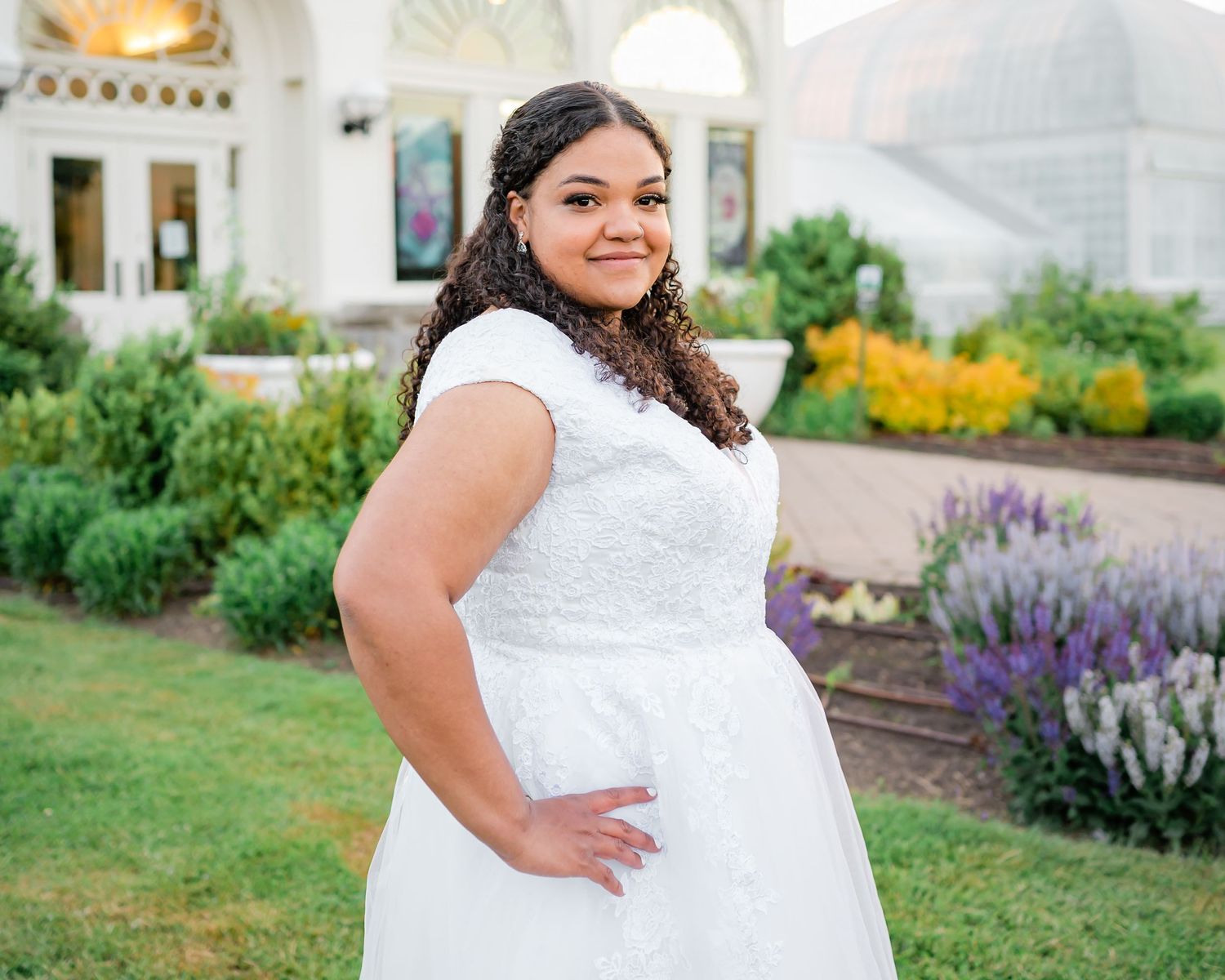 A bride in a white lace wedding dress poses in front of a garden with purple flowers.